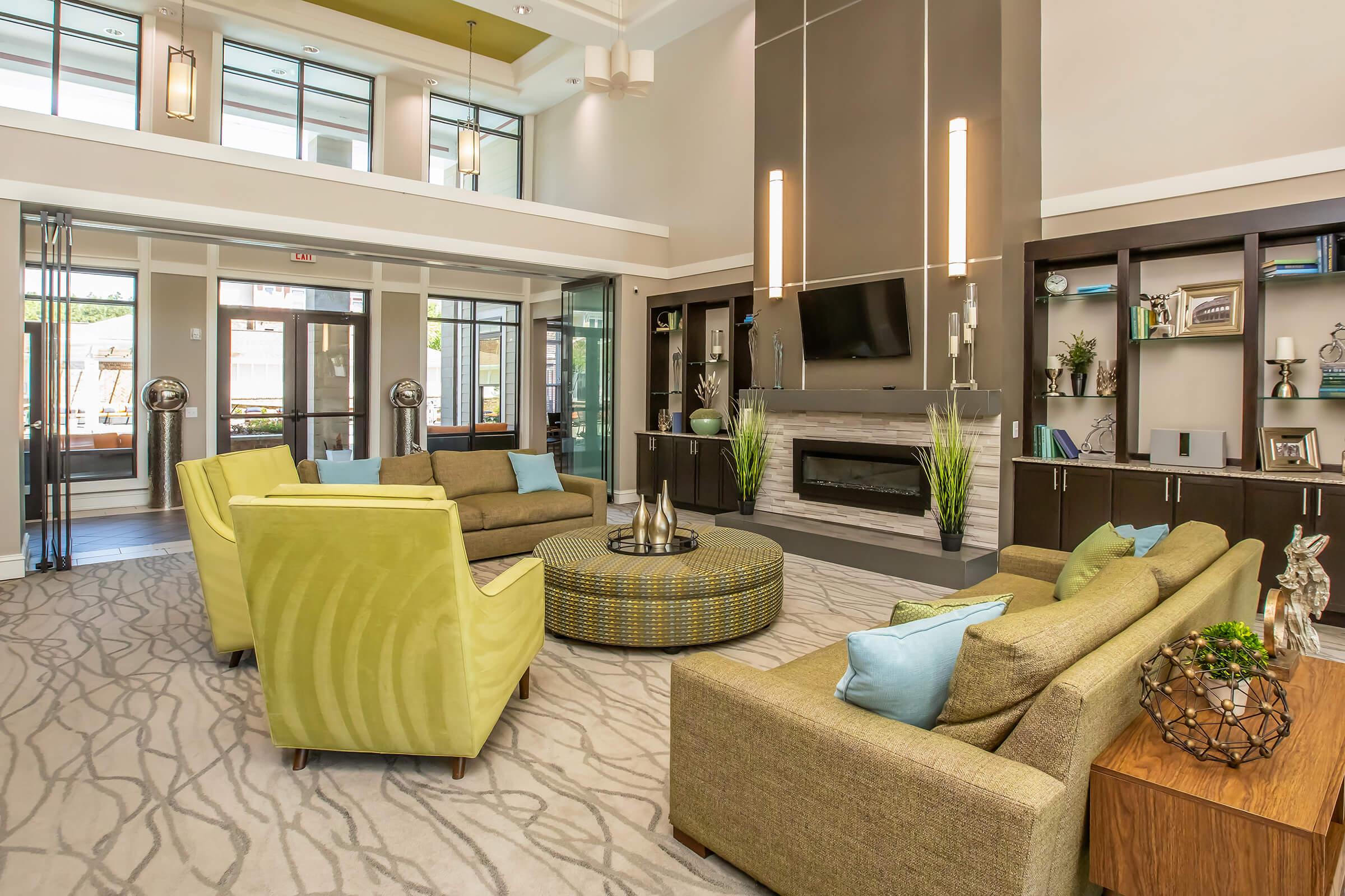 A modern and spacious lounge area featuring comfortable green and brown sofas, a circular accent table, and decorative plants. The room has large windows allowing natural light, a stylish fireplace, and contemporary lighting fixtures, creating a welcoming atmosphere for relaxation or socializing.