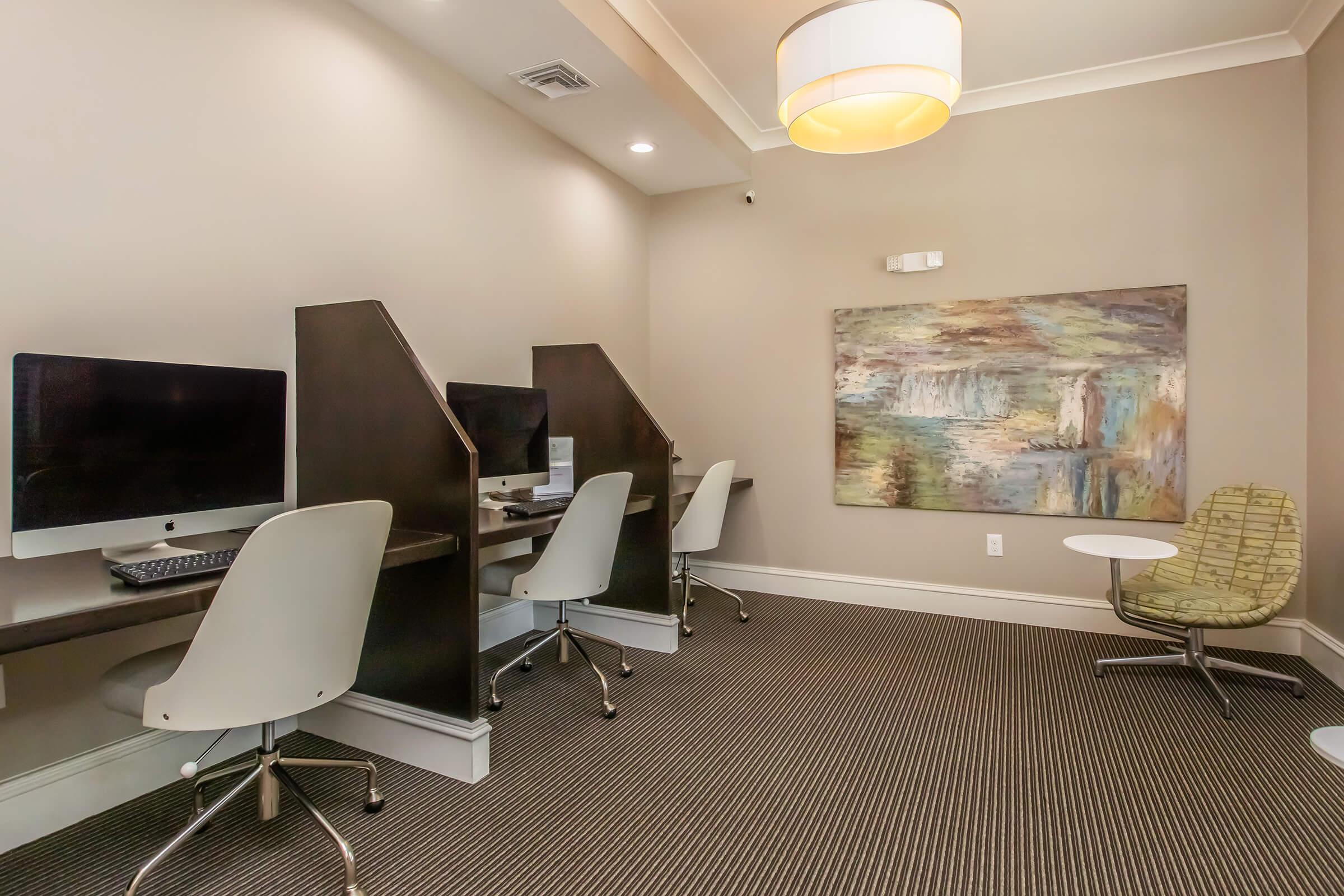 A modern workspace featuring three computer stations with privacy screens. The room has a neutral color palette with a decorative wall art piece, a round table, and a green chair, creating a professional and inviting atmosphere. Soft lighting enhances the overall ambiance.