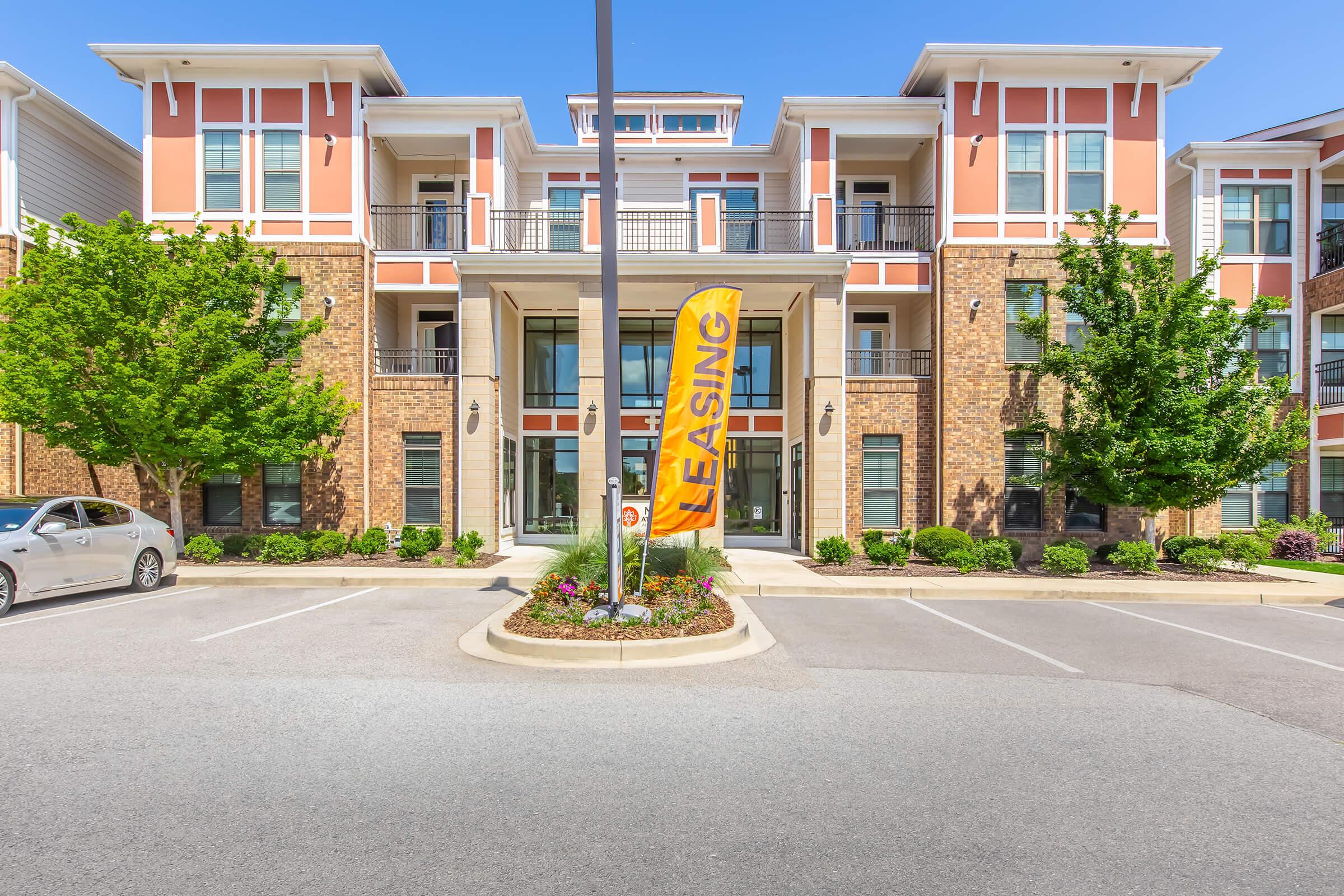 A modern apartment complex featuring a brick facade, large windows, and a leasing sign in front. The entrance is flanked by well-maintained landscaping, with small shrubs and flowers. Several parking spaces are visible in front of the building under a clear blue sky.