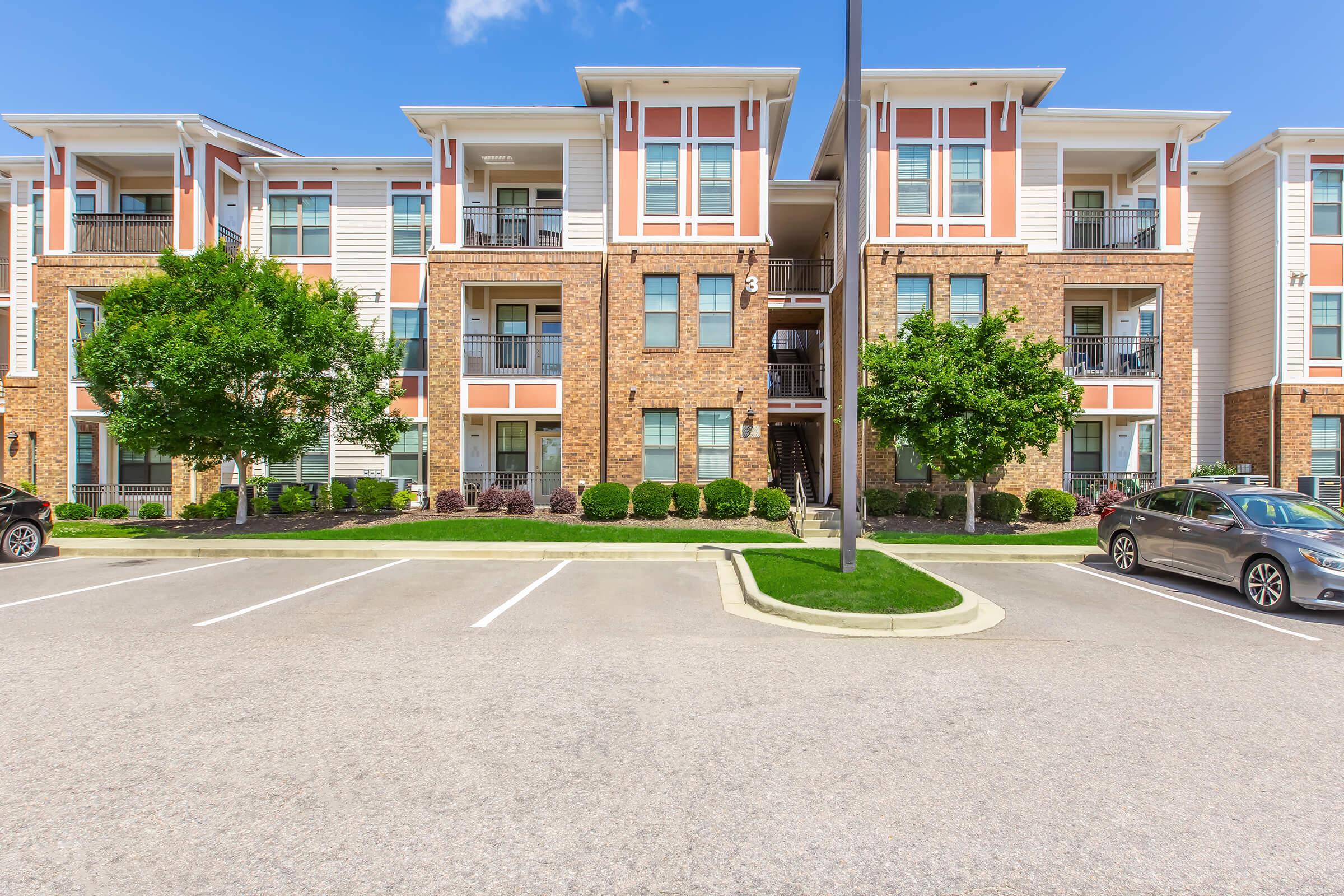 Modern apartment building with multiple balconies and large windows, surrounded by well-manicured shrubs and trees. The parking lot in front features several empty spaces, and the clear blue sky above adds to the bright and inviting atmosphere of the property.
