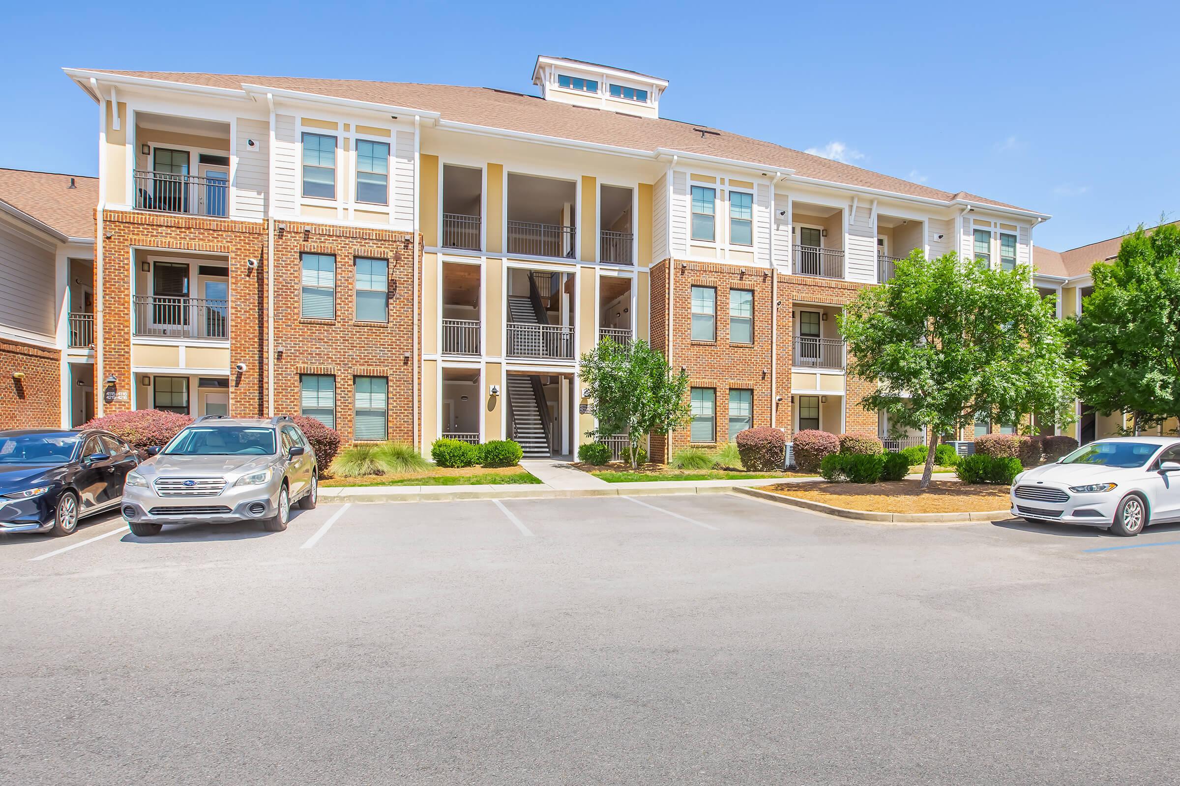 A modern brick apartment building with multiple balconies and three stories, surrounded by greenery. The parking lot in front contains several parked cars. The sky is clear and blue, indicating a sunny day.