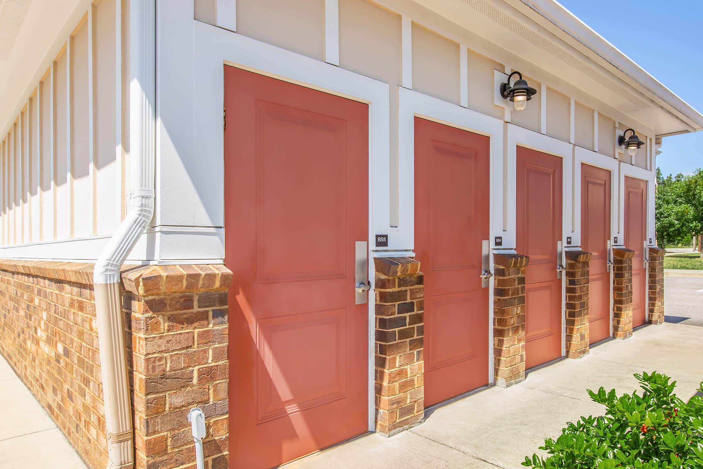A row of four orange doors set in a light-colored building with a brick foundation. Each door features a silver handle and a small identification plaque. The surrounding area is landscaped with green bushes and plants, under a clear blue sky.