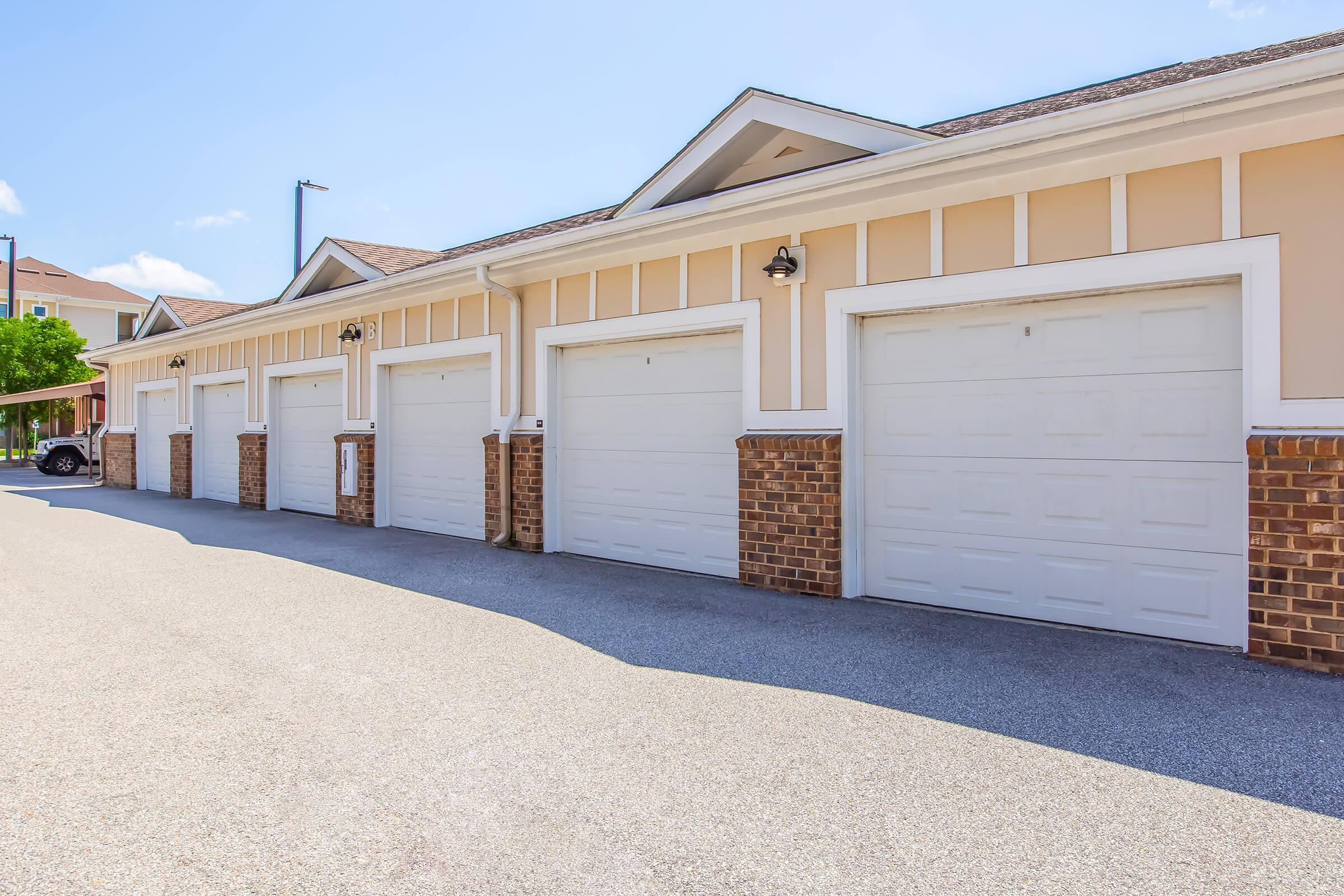 A line of closed garage doors in a residential setting, featuring beige siding and brick accents. The scene is under a clear blue sky, with a smooth, light-colored driveway in front. The garages are uniform in design, providing an organized appearance to the area.