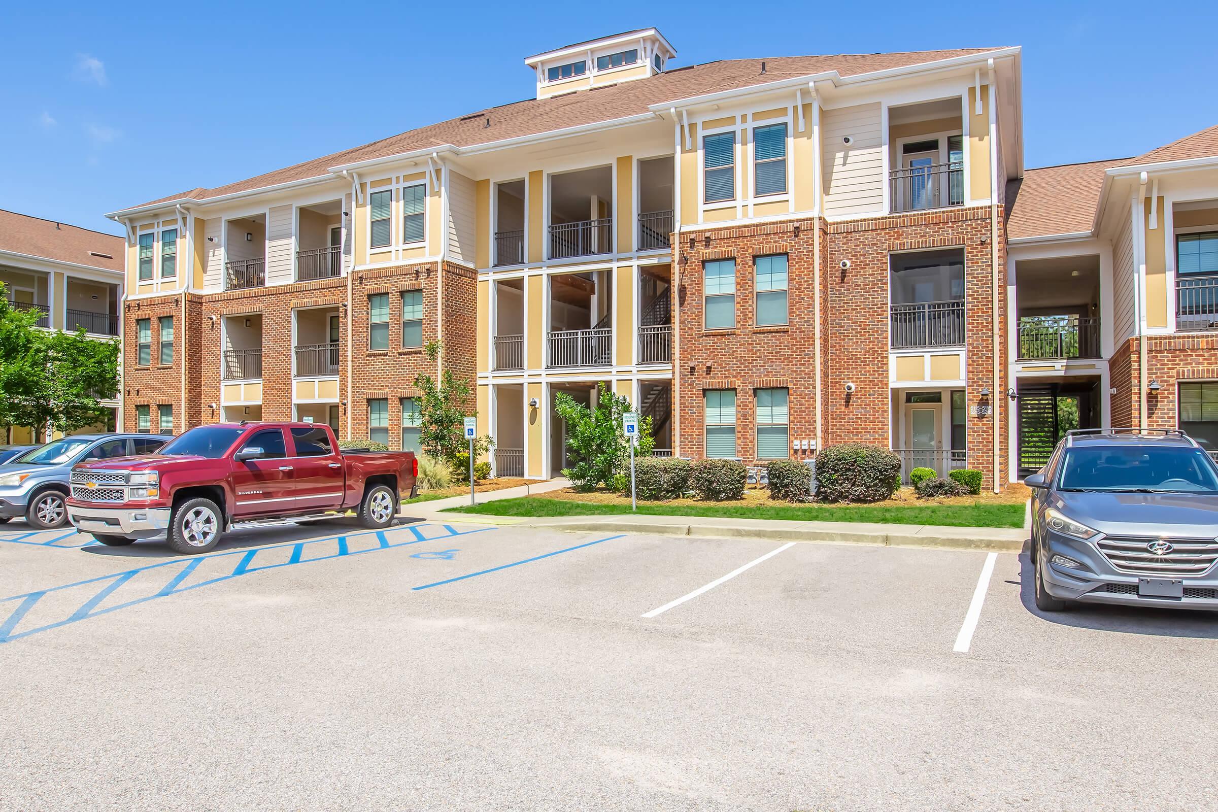 A sunny view of a multi-story apartment building with brick and siding exterior. The foreground features a parking area with several cars, including a red pickup truck and a silver sedan. Green shrubs and trees add some landscaping around the property.