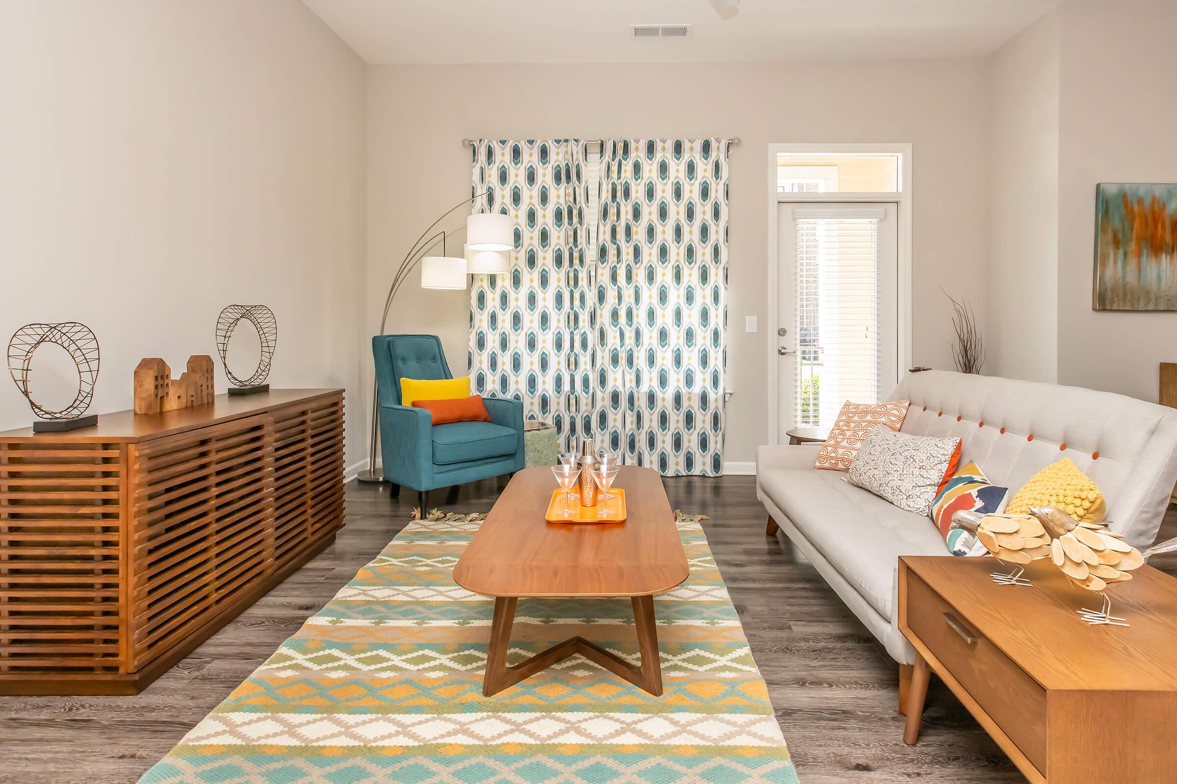 A modern living room featuring a light gray sofa with colorful pillows, a wooden coffee table, and a blue accent chair. The floor is covered with a geometric patterned rug, and there are decorative elements like vases and sculptures on display. Natural light streams in through a window with patterned curtains.