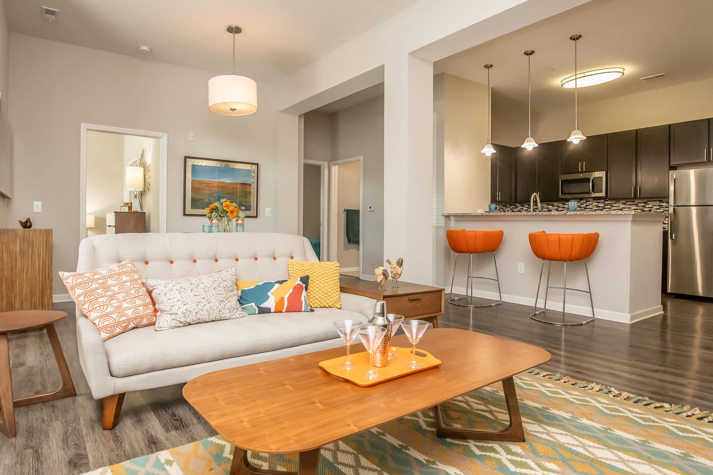 A modern living room featuring a light-colored sofa with colorful pillows, a wooden coffee table, and a vibrant area rug. In the background, there is a kitchen with dark wood cabinets and stainless steel appliances, along with stylish orange barstools, creating a warm and inviting atmosphere.
