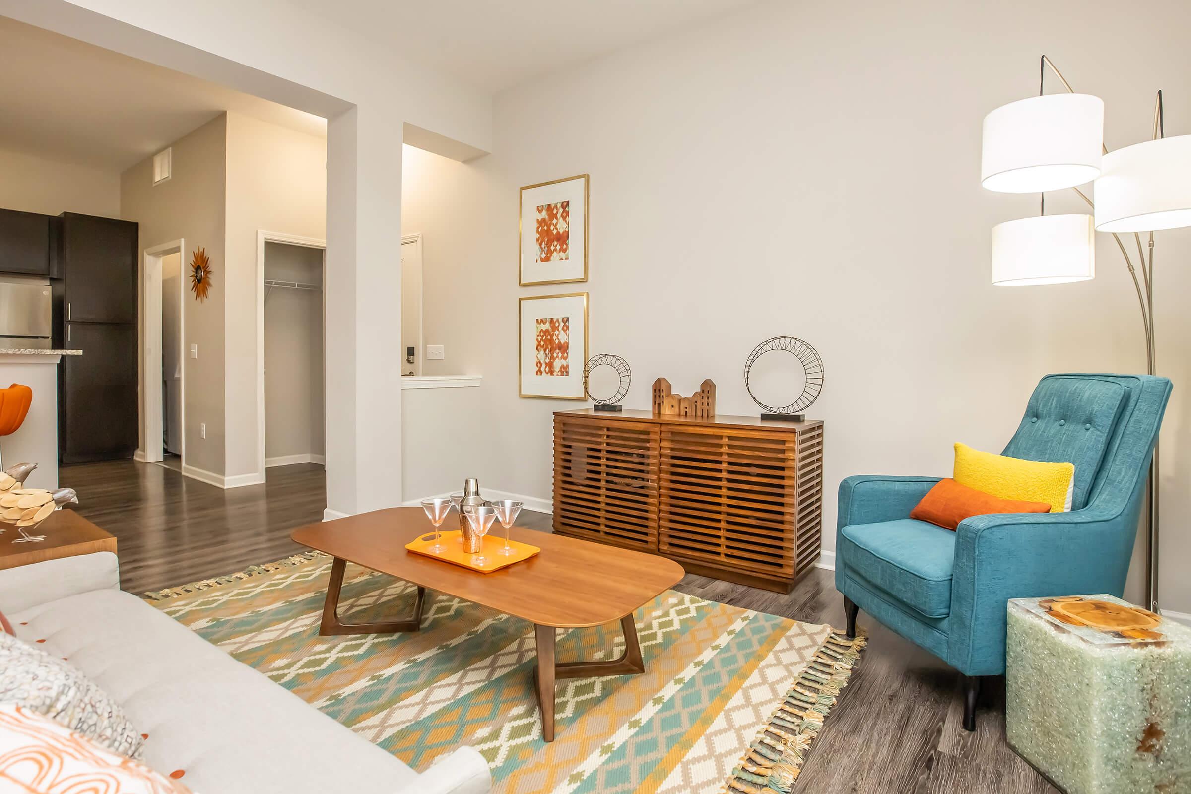 Cozy living room featuring a light-colored sofa, a wooden coffee table on a patterned rug, and an aqua-blue armchair with yellow cushions. A stylish lamp stands beside the chair, and art pieces decorate the walls. A wooden cabinet and open kitchen area are visible in the background.