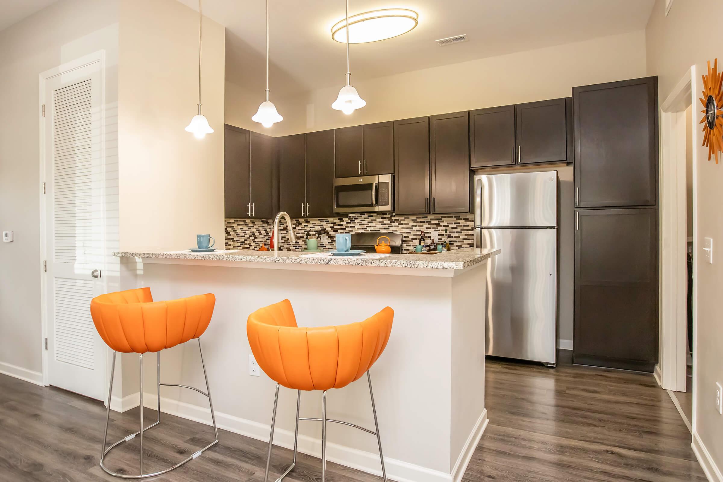 Modern kitchen with dark wood cabinets and stainless steel appliances. A granite countertop with two bright orange bar stools is in the foreground. Pendant lights hang above the counter, and a decorative wall clock is visible. The floor is a light wood finish, creating a warm atmosphere.