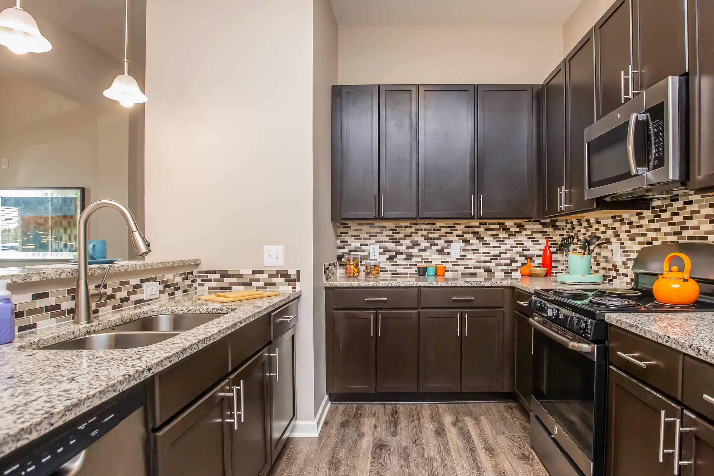 A modern kitchen featuring dark wooden cabinets, granite countertops, and a decorative backsplash. It includes stainless steel appliances, a double sink, and a gas stove. The kitchen is well-lit with pendant lighting and has various kitchen items such as pots and decorative objects on display.