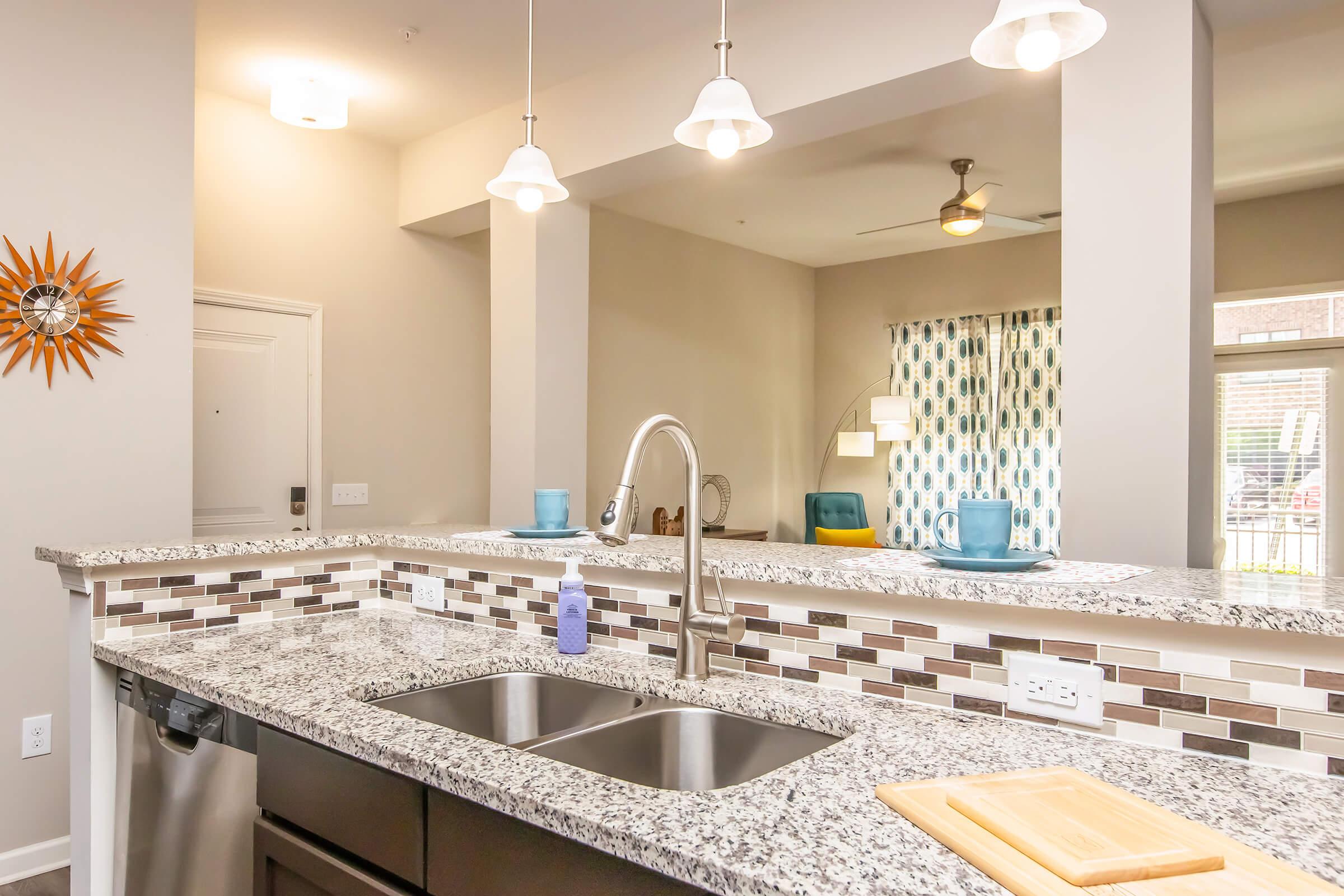 Modern kitchen with granite countertops and stainless steel double sinks, featuring pendant lights above, a backsplash of mosaic tiles, and a view into a bright living area with a ceiling fan. A wooden cutting board rests on the counter, and decorative elements enhance the space.
