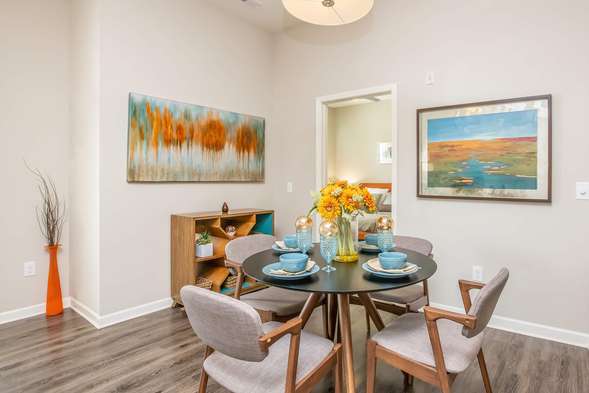 A modern dining area featuring a round black table set with blue dishes and a floral centerpiece. Surrounding the table are stylish wooden chairs. The walls are adorned with abstract artwork in soft colors, and there's a wooden shelf displaying decorative items. Hardwood flooring completes the contemporary look.