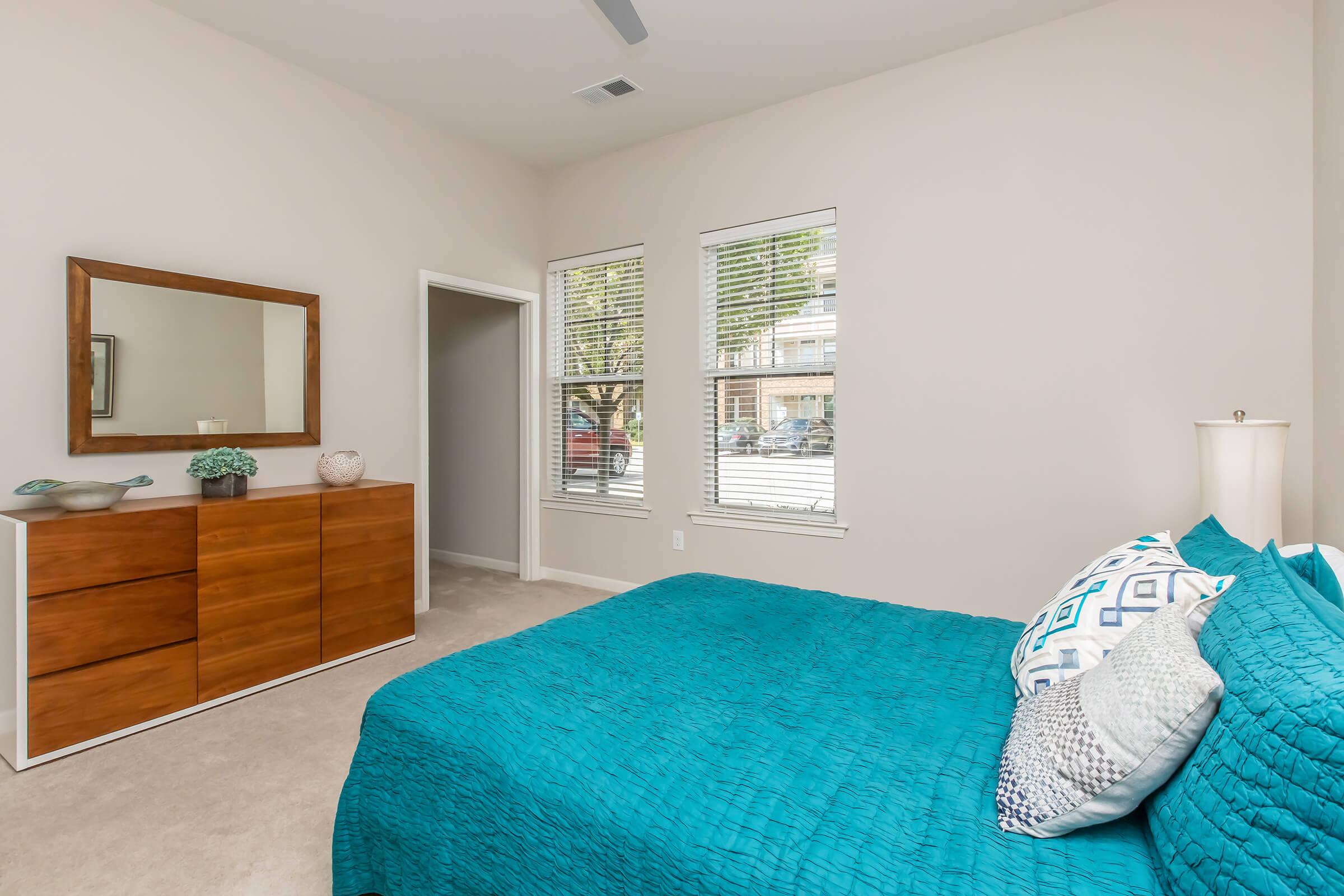 A brightly lit bedroom featuring a large window with white blinds. The room has a bed with a vibrant teal bedding set and decorative pillows. A modern wooden dresser is positioned against the wall, and a mirror above it reflects the space. Fresh greenery is displayed on the dresser, adding a pop of color.