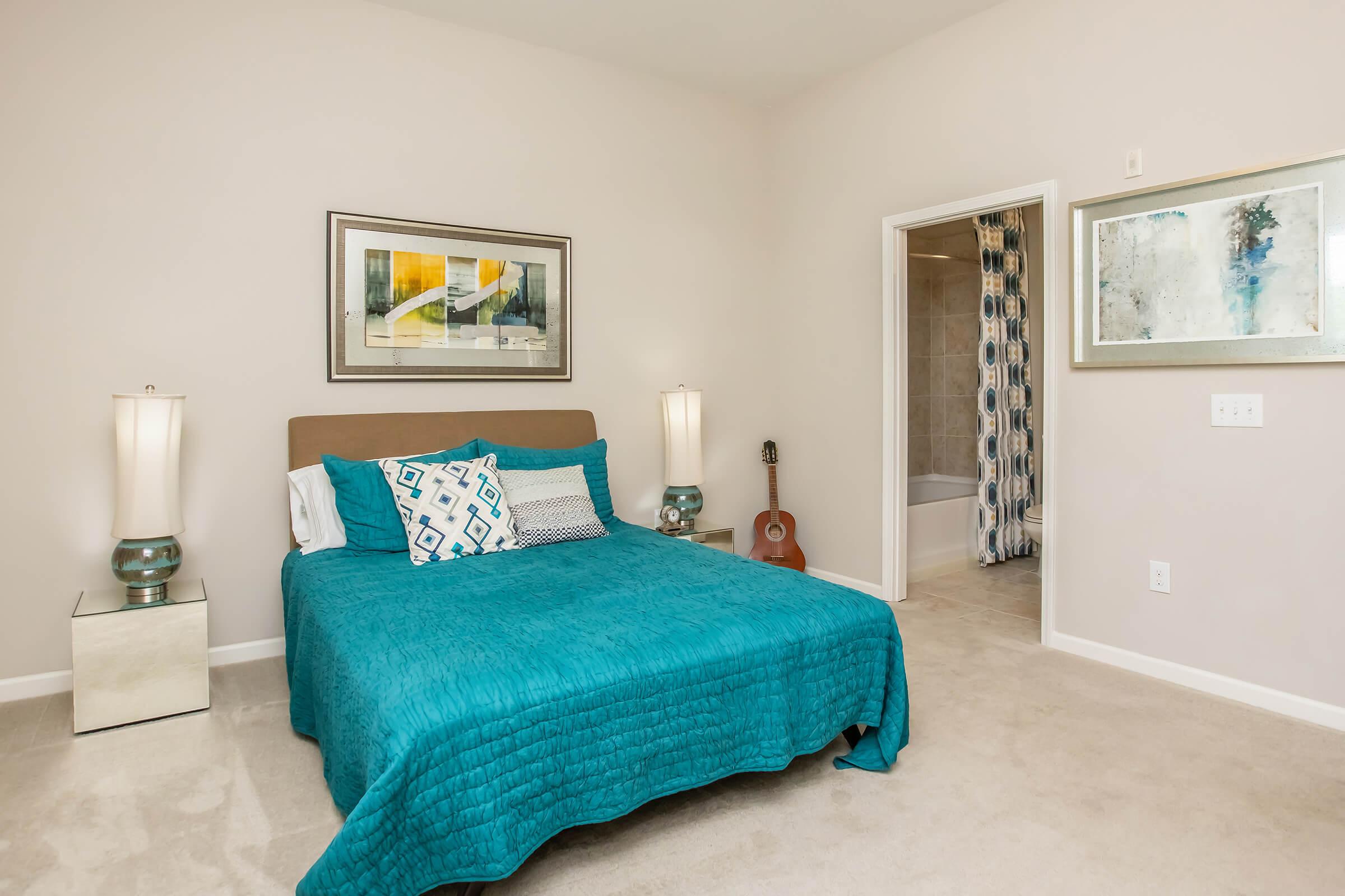 A well-lit bedroom featuring a queen-sized bed with a teal quilt and decorative pillows. There are bedside lamps on mirrored tables, a guitar leaning against the wall, and a doorway leading to a bathroom. Abstract artwork adorns the walls, enhancing the contemporary decor. The carpeted floor adds warmth to the space.