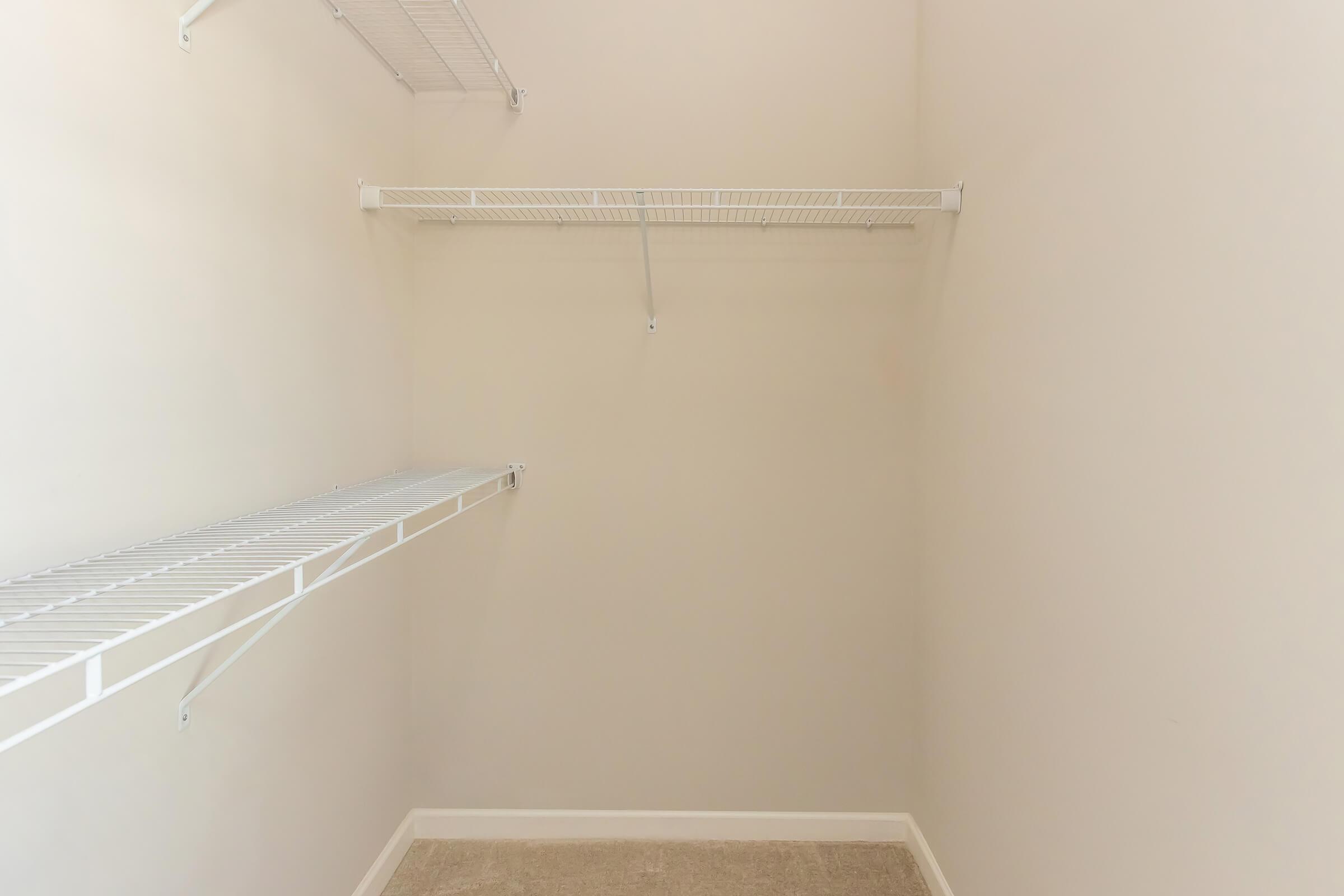 An empty closet with white wire shelving on the left and right walls, featuring a light beige wall and carpeted floor. The shelves are bare, creating a spacious and minimalist appearance.