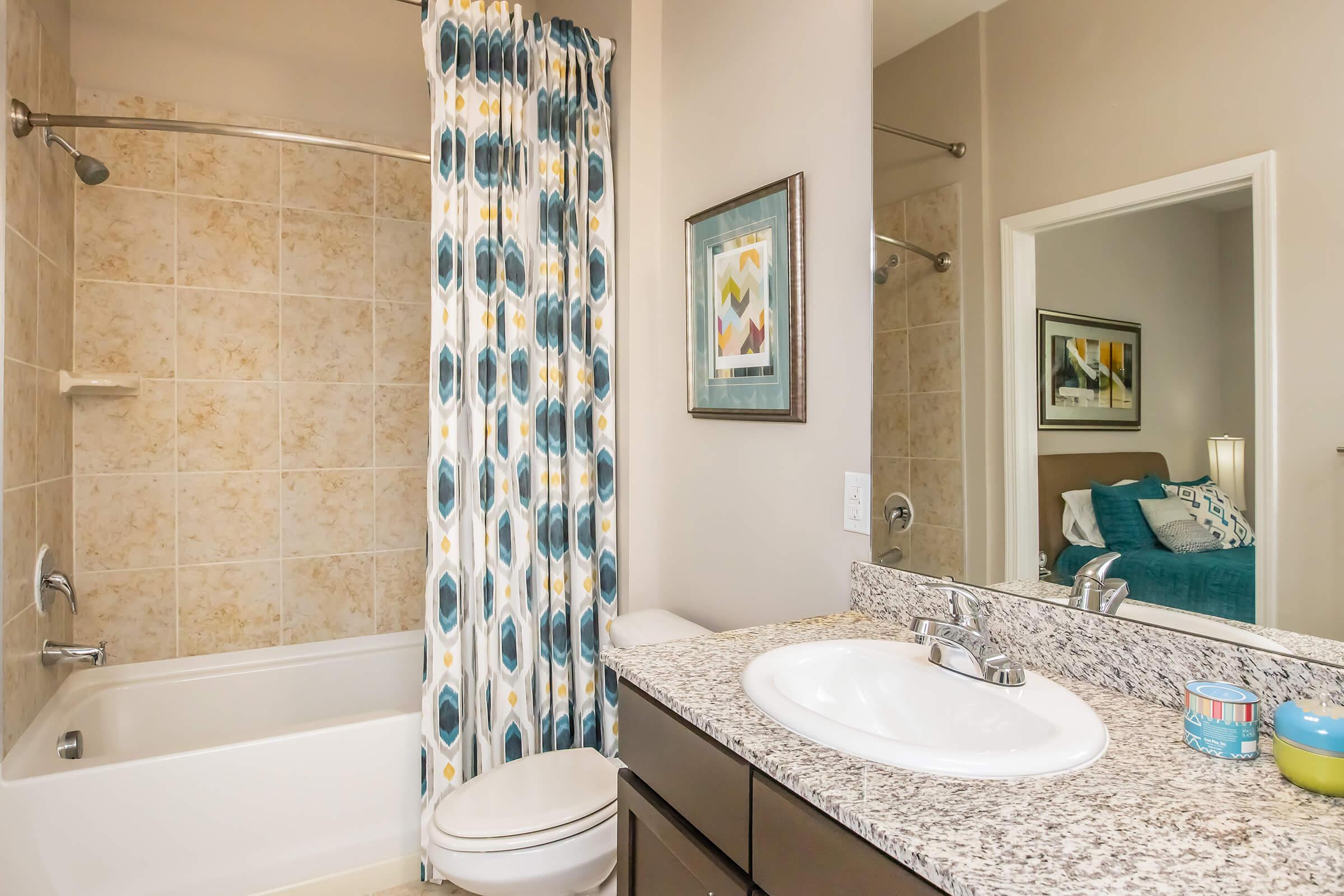 A stylish bathroom featuring a bathtub and shower combination with a light-colored tile backdrop. There's a modern sink with a granite countertop and a mirror above. The space is accented by a decorative shower curtain and artwork on the walls, creating a clean and inviting atmosphere.