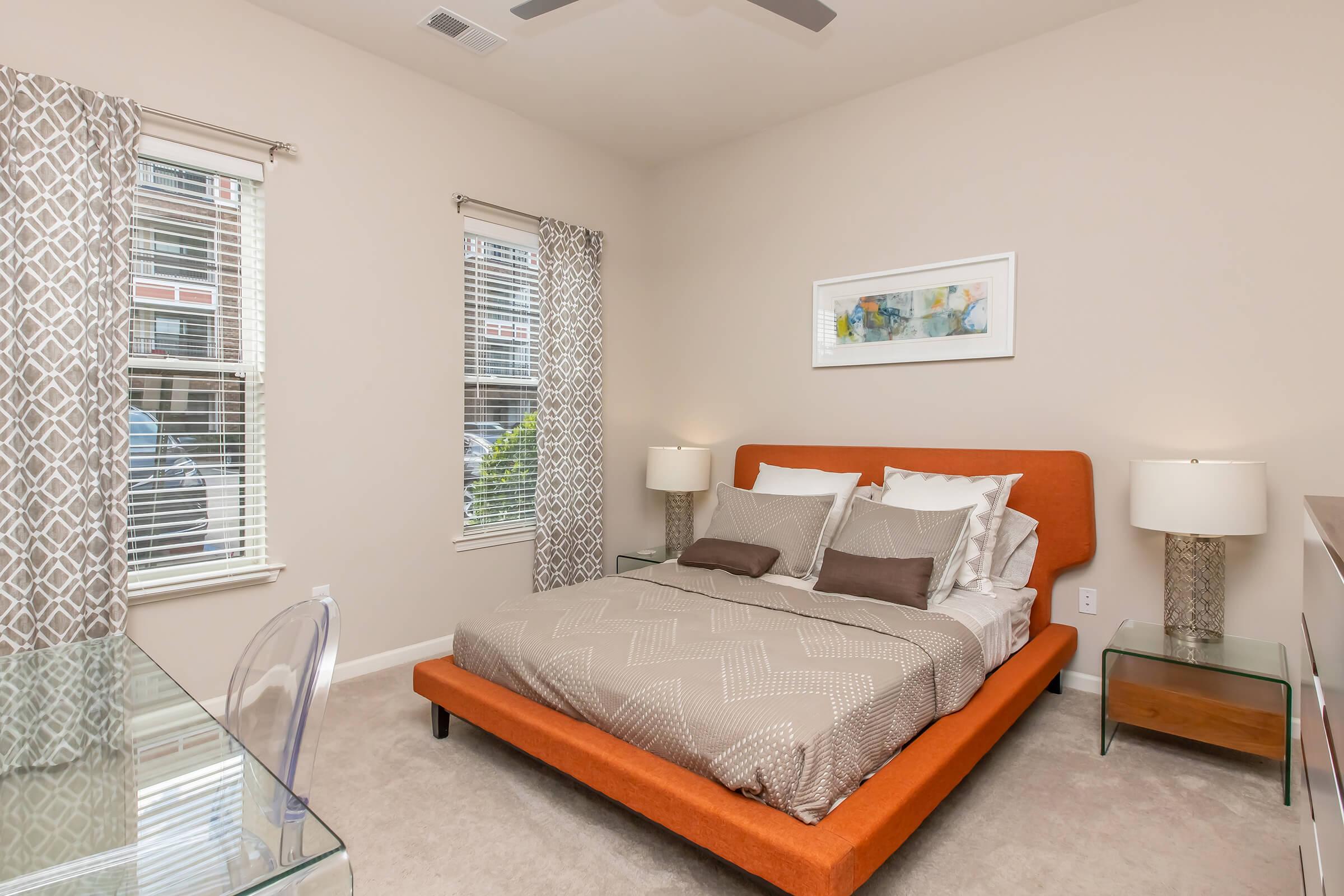 A modern bedroom featuring an orange upholstered bed with decorative pillows, two bedside lamps on glass nightstands, a glass desk, and large windows with white curtains. The walls are painted in a soft neutral color, contributing to a cozy and inviting atmosphere.