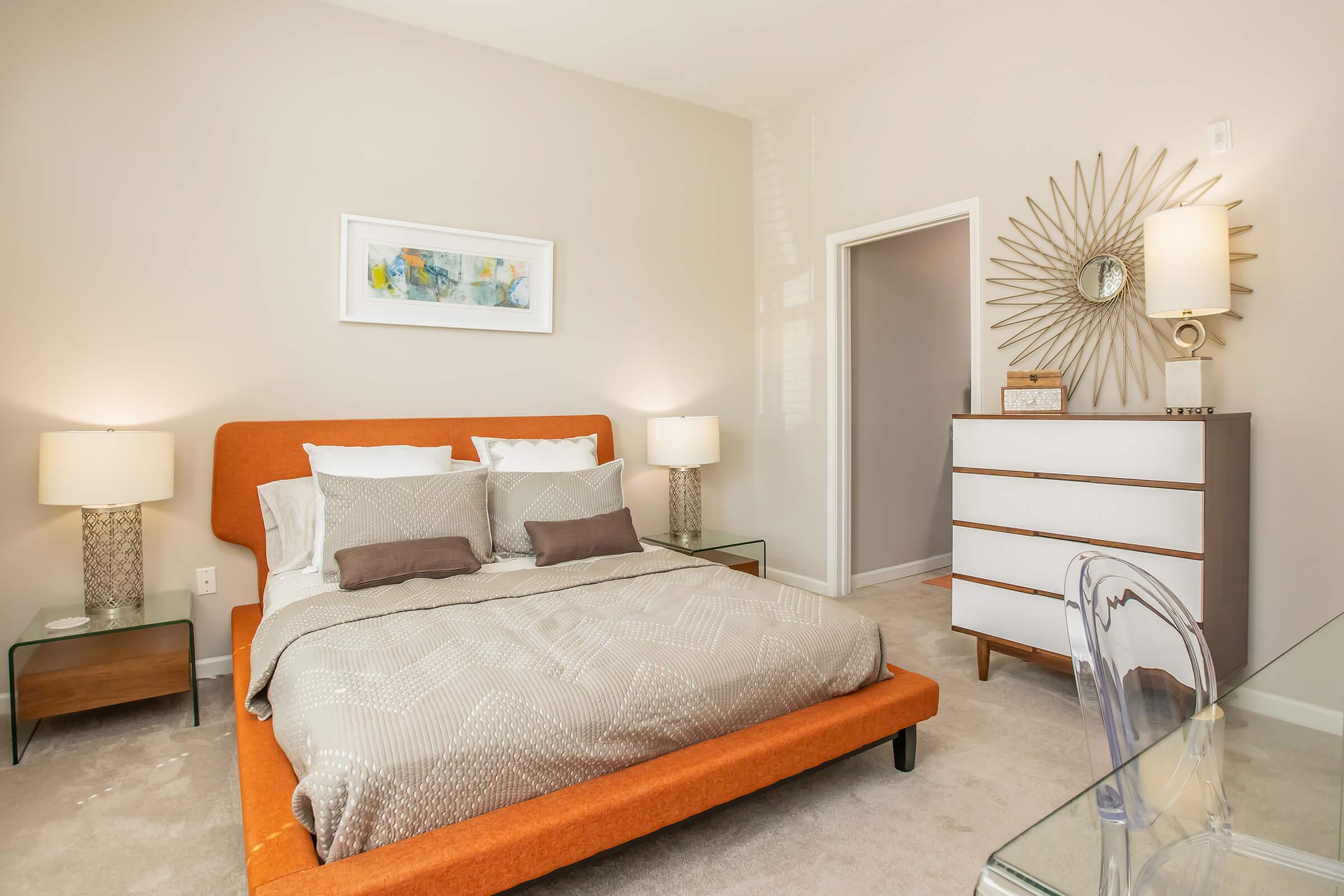 A modern bedroom featuring an orange upholstered bed with layered bedding, two bedside tables with lamps, a stylish dresser, and a large mirror. The decor includes a colorful wall art piece and a sunburst mirror, creating a warm and inviting atmosphere. Soft beige walls and carpet enhance the cozy feel.