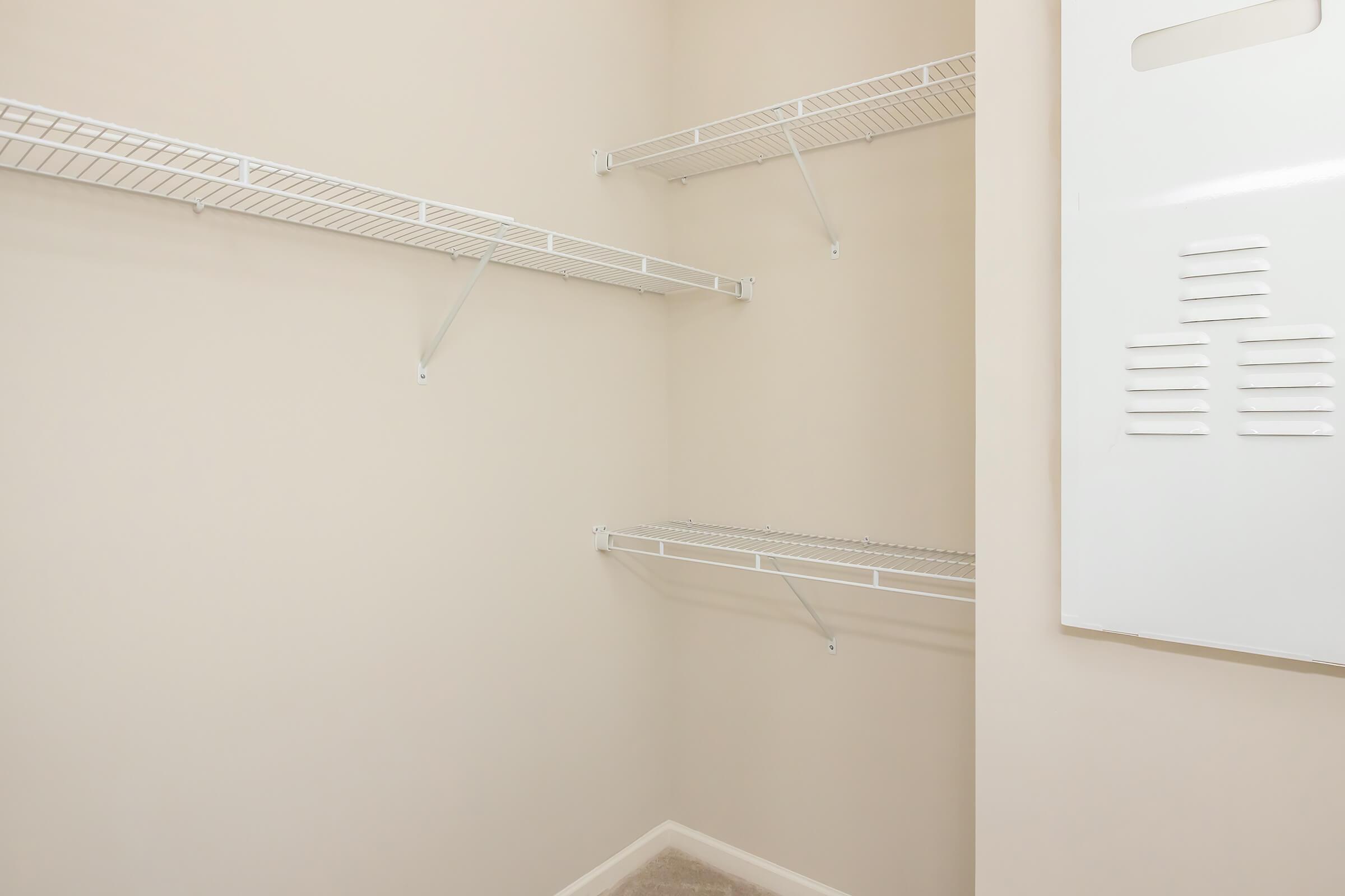 Empty closet features beige walls and two wire shelves mounted on one side. A blank white panel is mounted on the adjacent wall. The floor is carpeted, and the space is well-lit, creating a clean, organized appearance.