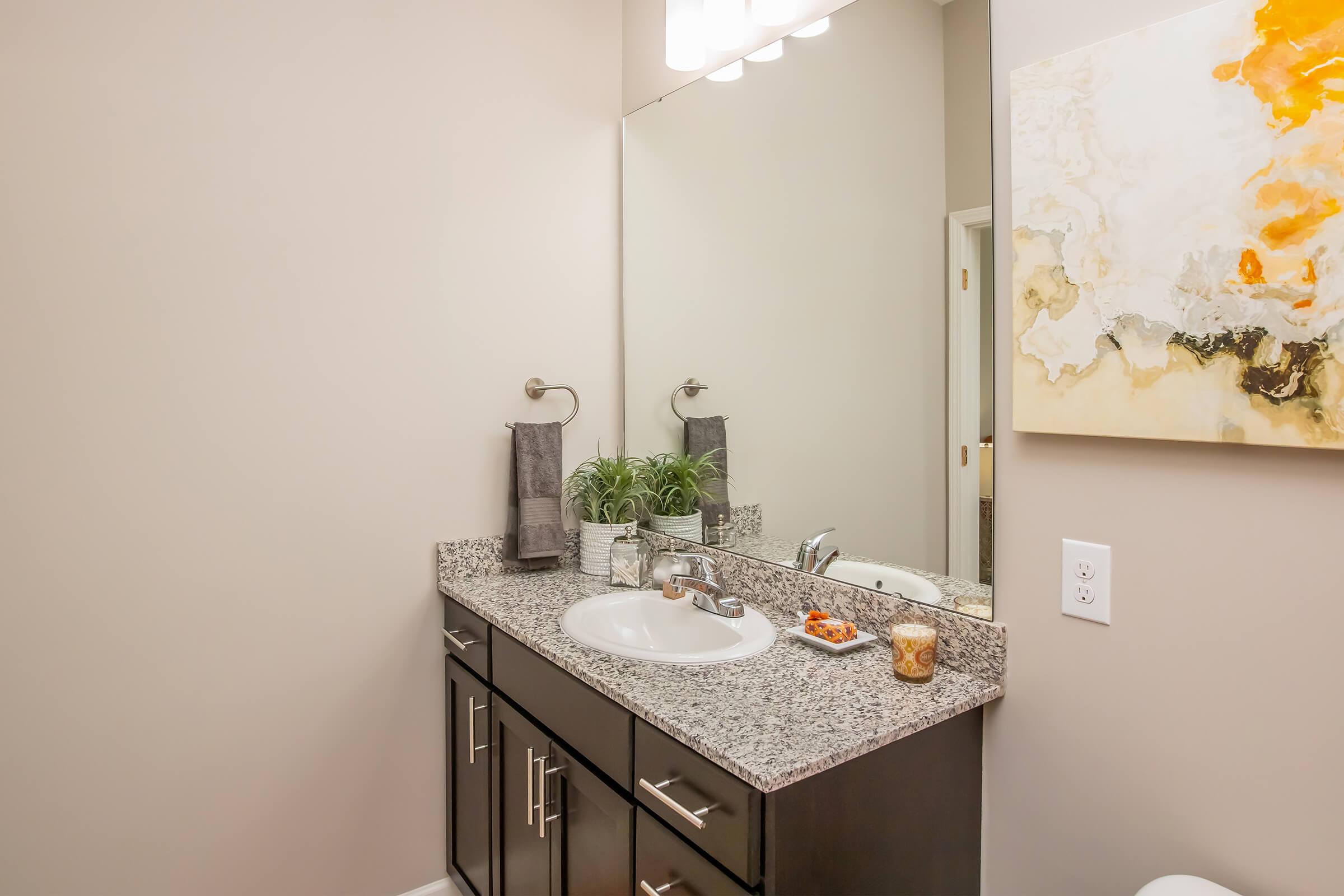 A modern bathroom featuring a dark wood vanity with a granite countertop, a round sink, and a large mirror. On the counter, there are hand towels, a small plant, a candle, and a decorative dish. The wall opposite has an abstract artwork with yellow and gray tones. Soft lighting enhances the clean and stylish atmosphere.