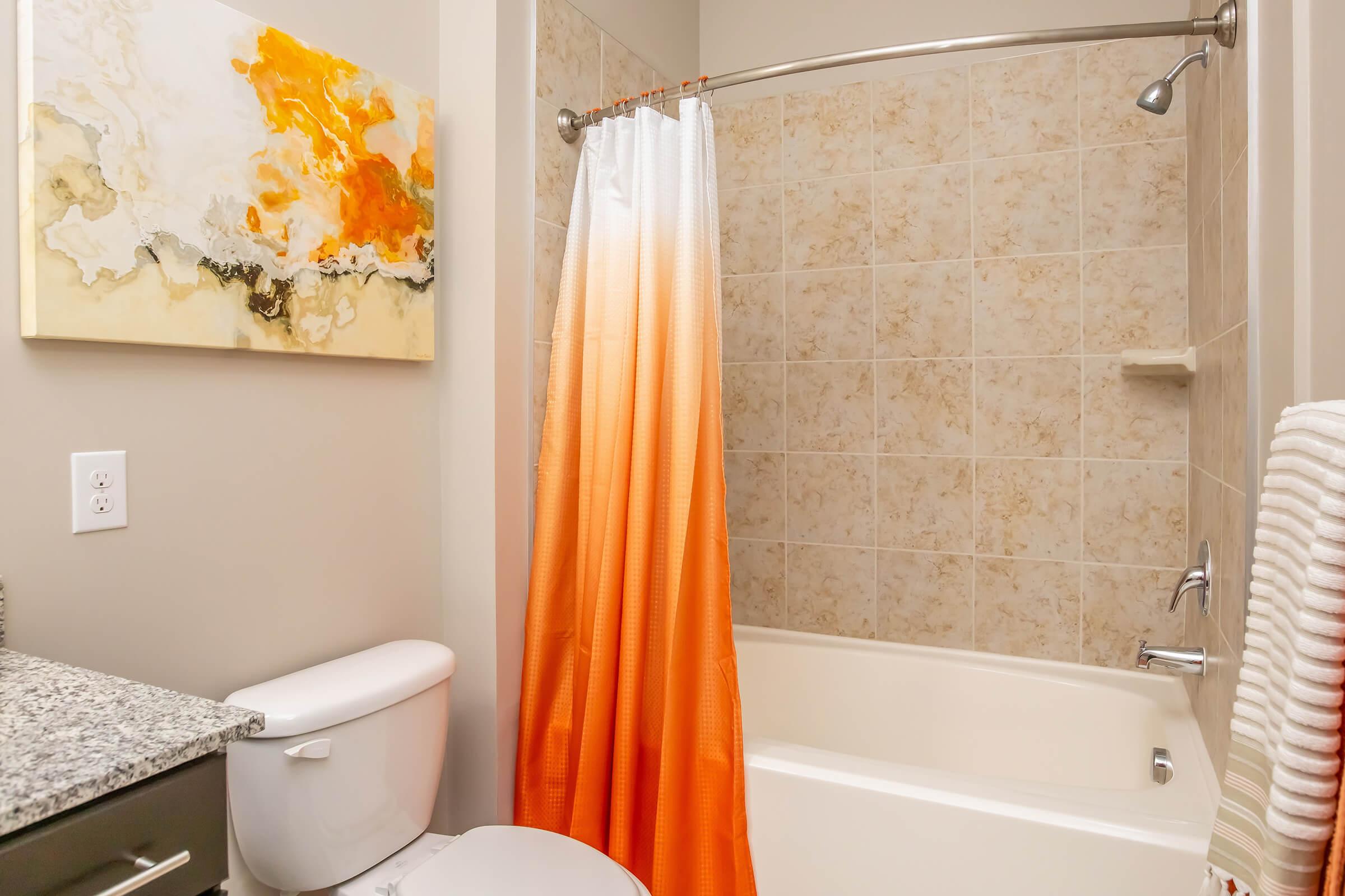 A modern bathroom featuring a white bathtub with an orange gradient shower curtain, a compact toilet, and a granite countertop. The wall has abstract artwork in orange and cream tones. The tiles are light-colored, creating a bright and clean atmosphere. Soft towels hang nearby, adding to the decor.
