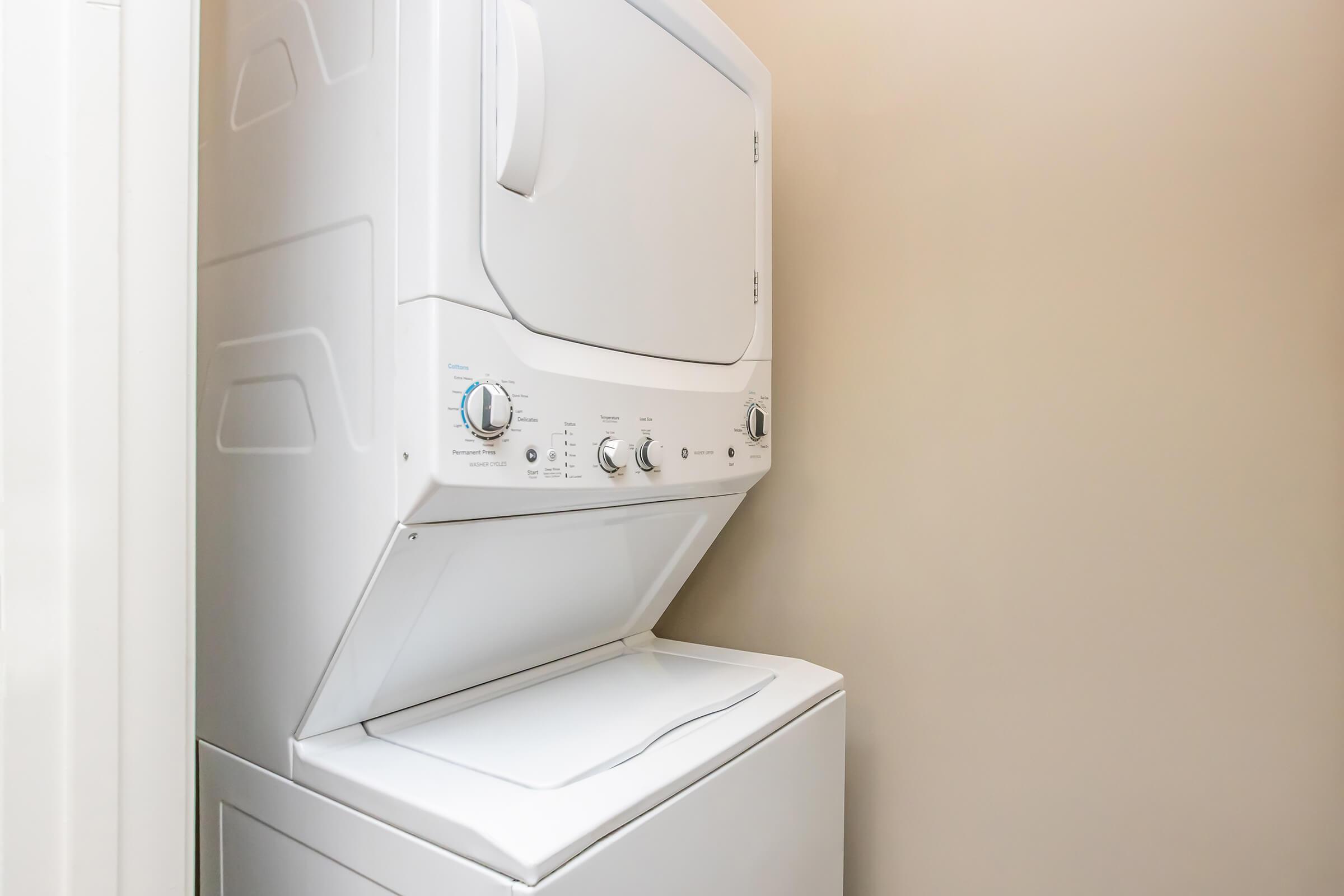 Stacked washer and dryer units in a laundry area, set against a beige wall. The washer is at the bottom and the dryer is on top, featuring control knobs and a door. The space is organized and functional, emphasizing a compact design for home laundry needs.