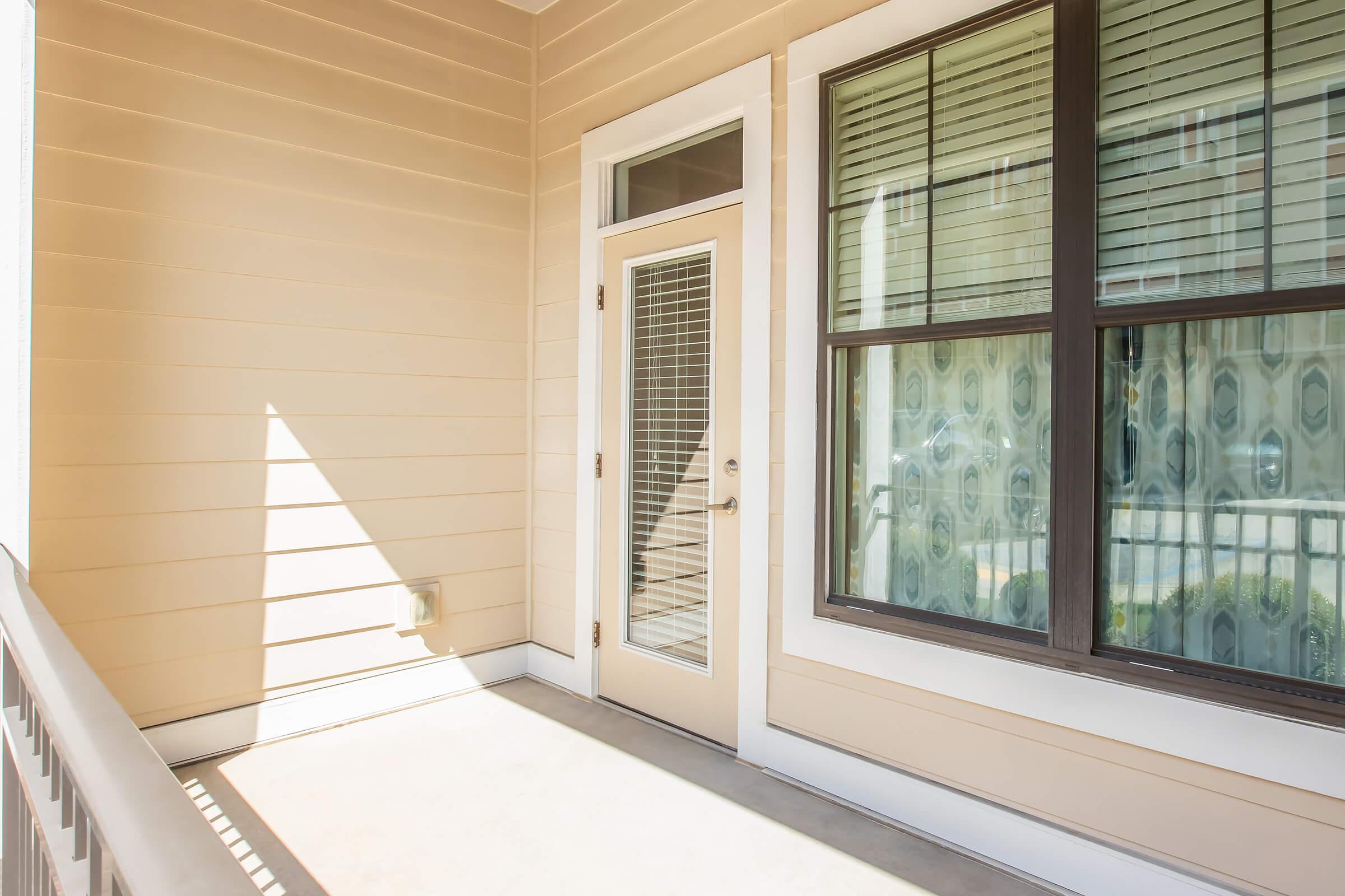 A bright and spacious balcony area with beige walls. There is a glass-paneled door leading inside, alongside a large window with dark frames and a patterned curtain. The floor is concrete, and sunlight creates a warm atmosphere in this outdoor space.