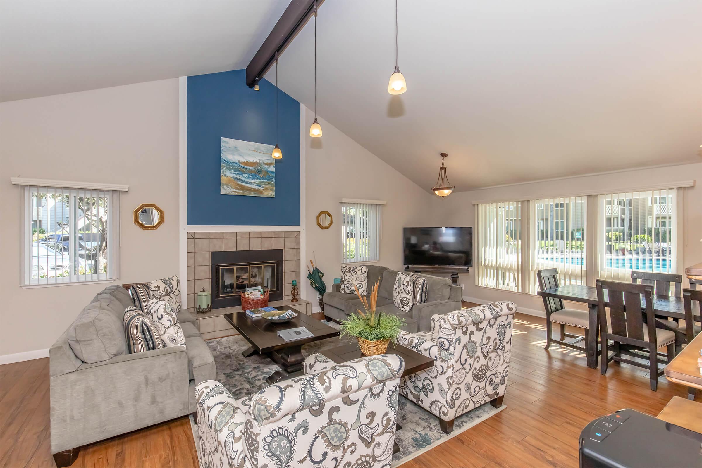 Spacious living room featuring a decorative fireplace, comfortable sofas with intricate patterns, a coffee table, and a flat-screen TV. Large windows provide natural light, and a dining area with a wooden table is visible in the background. The walls are painted in neutral tones with an accent blue wall.