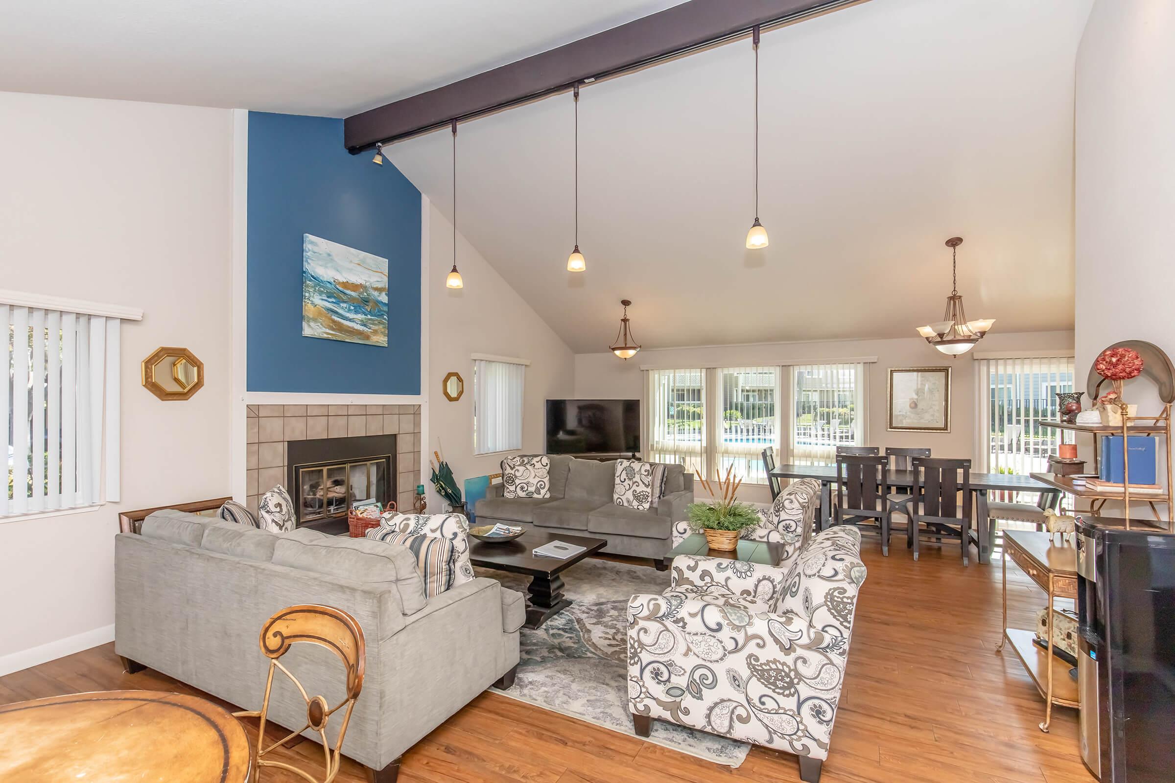 A spacious living room with high ceilings, featuring a gray sectional sofa, patterned armchairs, and a decorative area rug. A coffee table sits in the center, and a fireplace is visible on one side. Natural light floods the space through large windows, with a dining area and a television in the background.