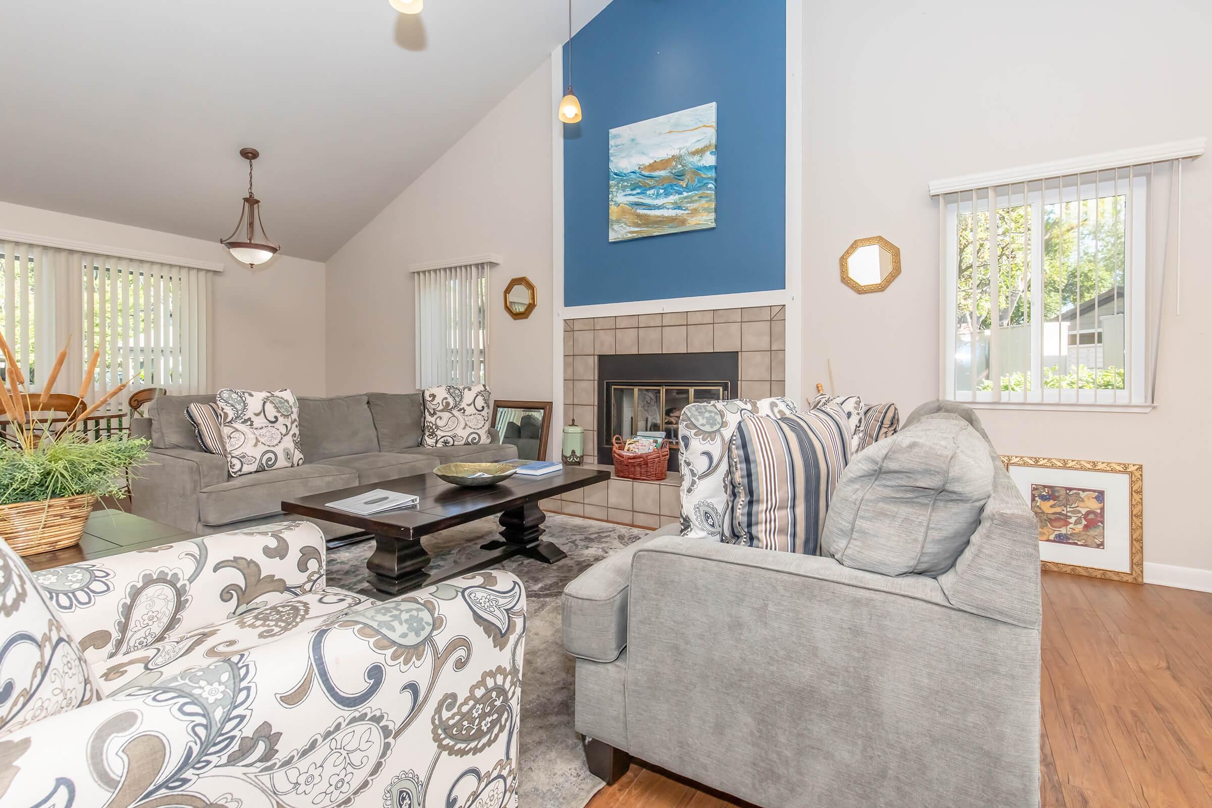 A cozy living room featuring a combination of gray sofas and patterned chairs, with a coffee table at the center. There's a fireplace with a decorative painting above it, and sunlight streams in through large windows adorned with blinds. A decorative basket and wall art enhance the inviting atmosphere.
