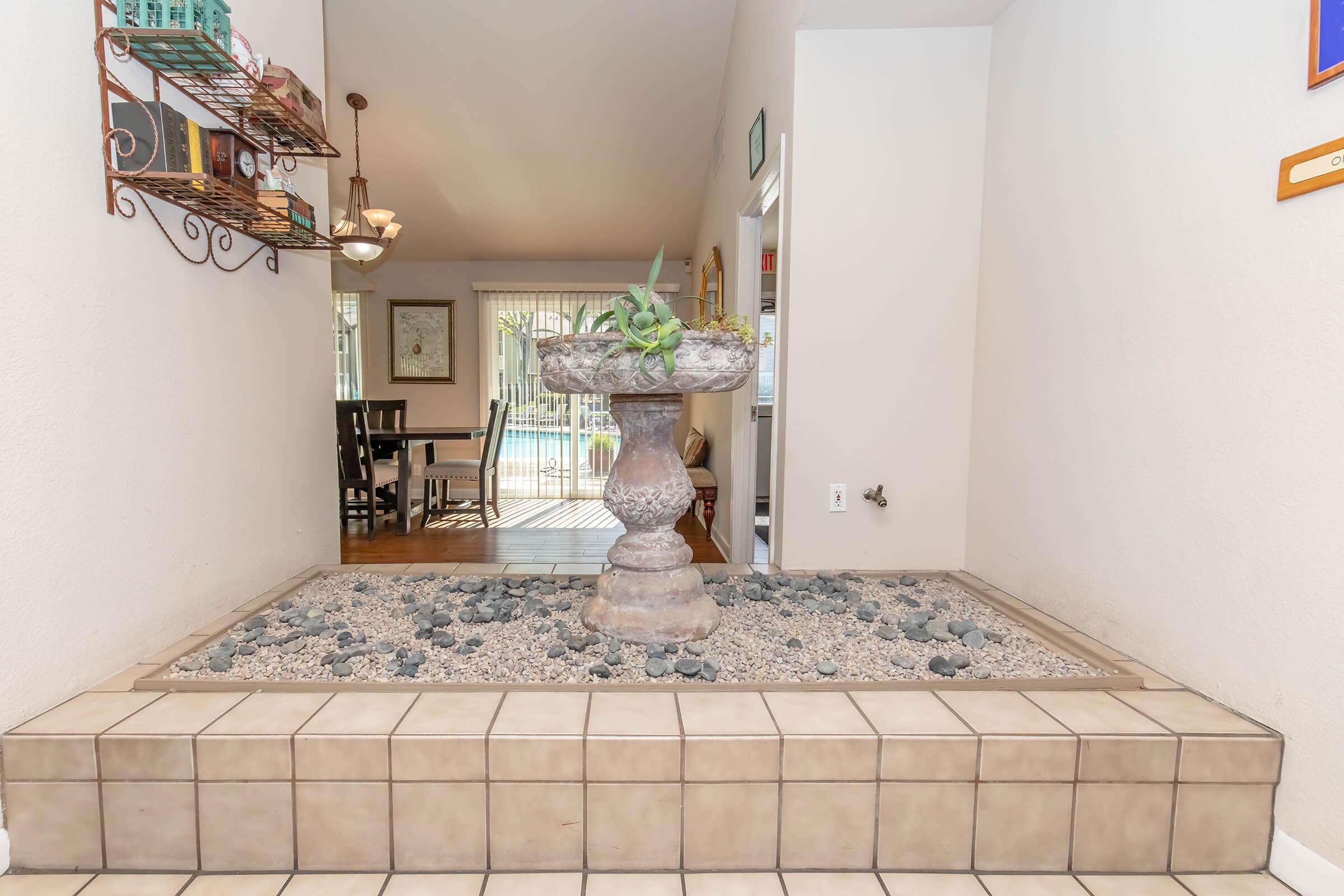 Indoor entryway featuring a decorative stone fountain surrounded by small rocks on a tiled surface, leading into a bright room with windows. The area is well-lit, with a hanging light fixture visible above.