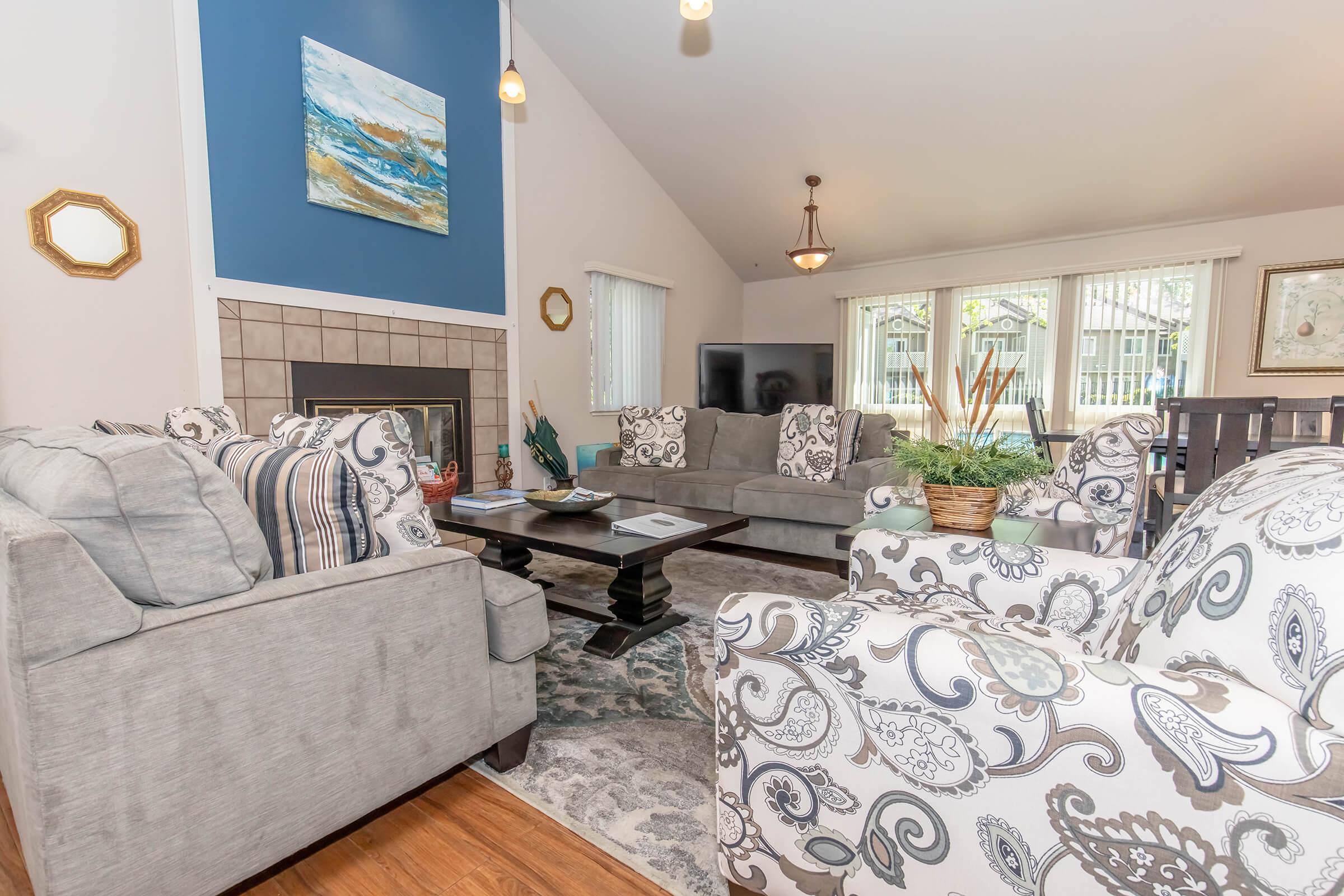 A cozy living room with a modern design features two patterned armchairs and a plush gray sofa arranged around a dark coffee table. The space is illuminated by pendant lights and has a light, airy feel, with a stone fireplace and large windows allowing natural light in. Decorative plants and artwork add a warm touch.