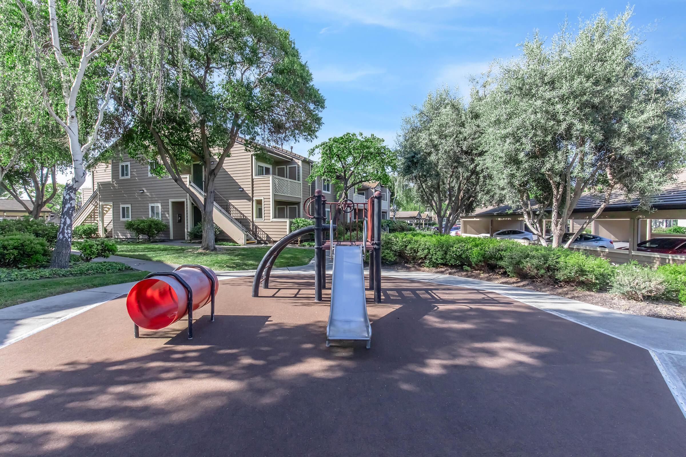 A children's playground featuring a red tunnel slide and climbing structure, surrounded by lush green trees and bushes. In the background, there are residential buildings and parked cars, all under a clear blue sky. The playground area is neatly maintained with a soft ground surface.