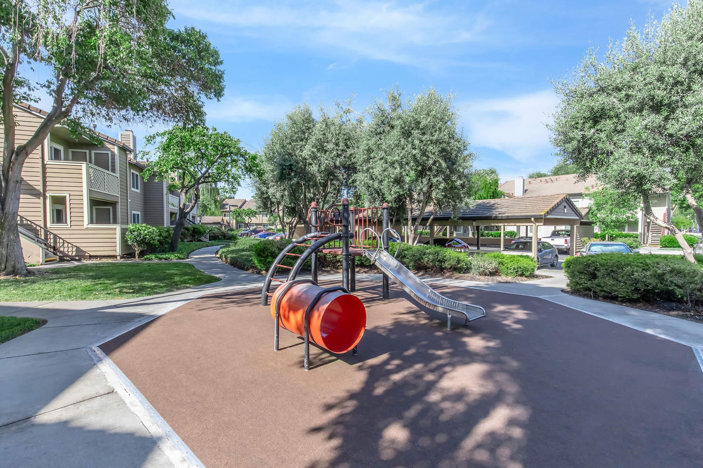 A playground featuring a slide and a tunnel, surrounded by green trees and grassy areas. Nearby apartment buildings are visible, along with a path leading through the park-like setting. The sky is clear and blue, adding to the outdoor ambiance.