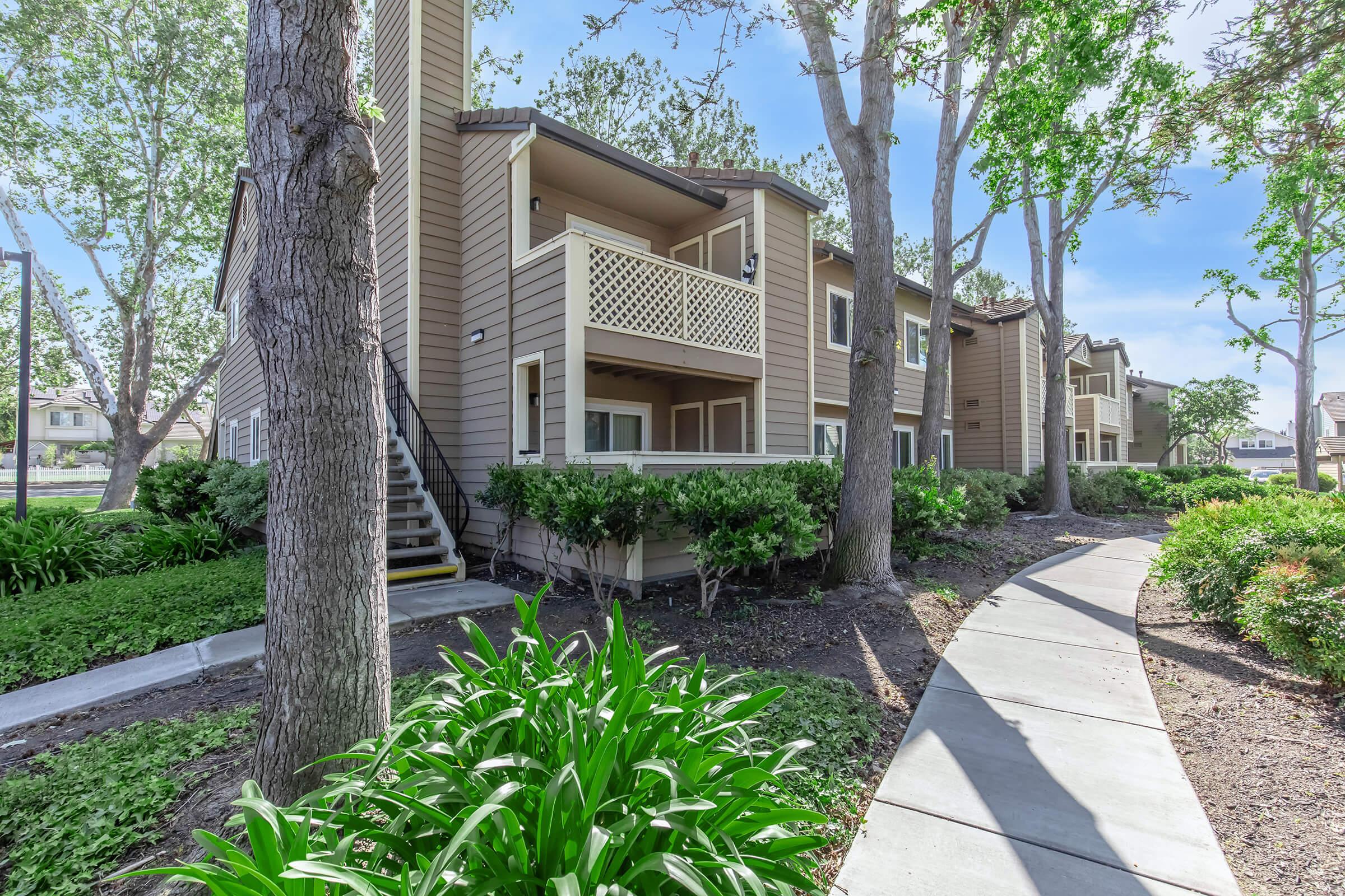 A view of a residential building with multiple balconies, surrounded by well-maintained gardens. The area features trees and a pathway leading towards the front entrance, creating a serene outdoor space. Bright blue sky in the background adds to the inviting atmosphere.