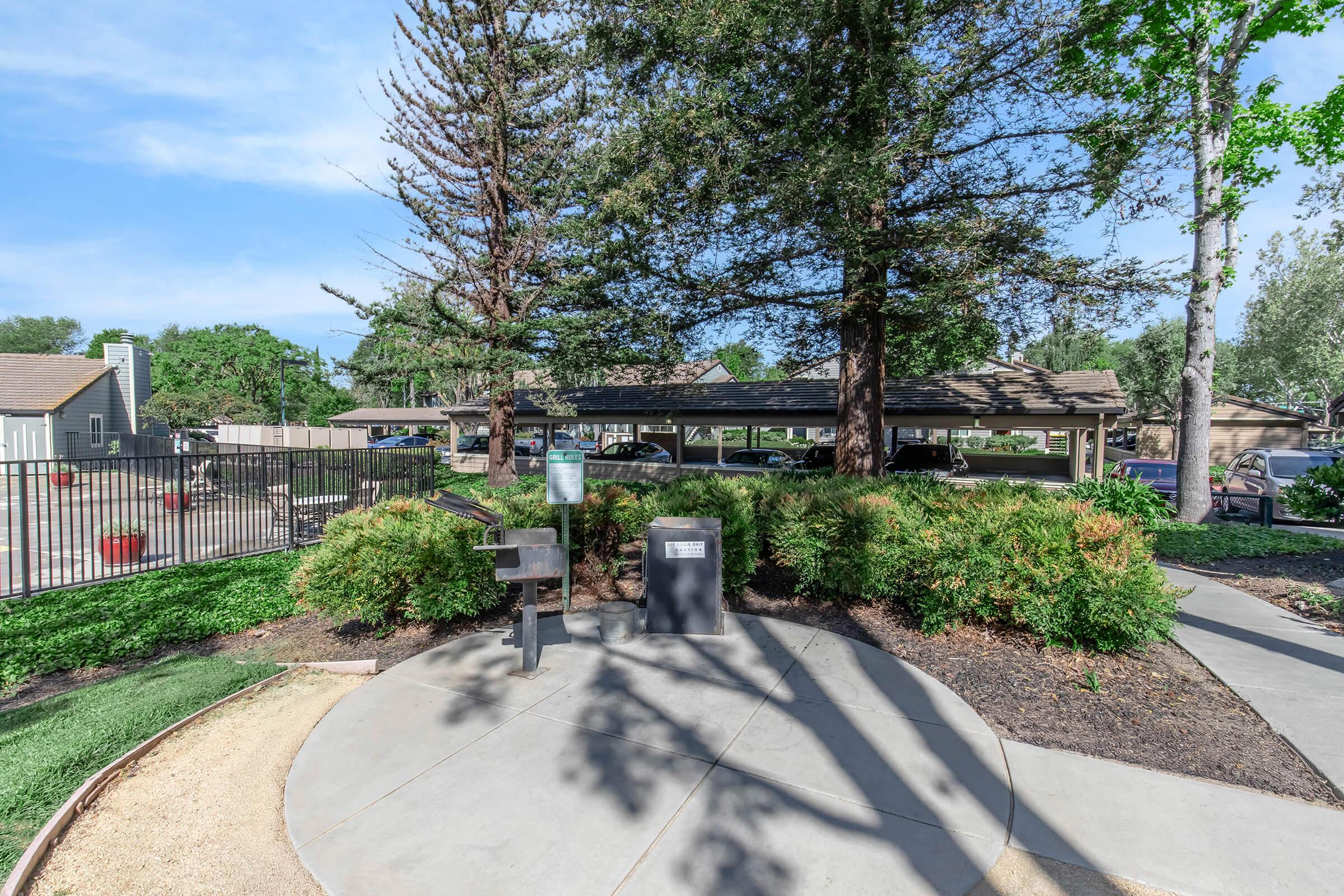 A landscaped area with a concrete pathway, featuring a grill and waste disposal station. Trees and shrubs surround the space, while a few cars are visible in the background near a covered parking area. A chain-link fence encloses the larger property, with clear blue skies above.