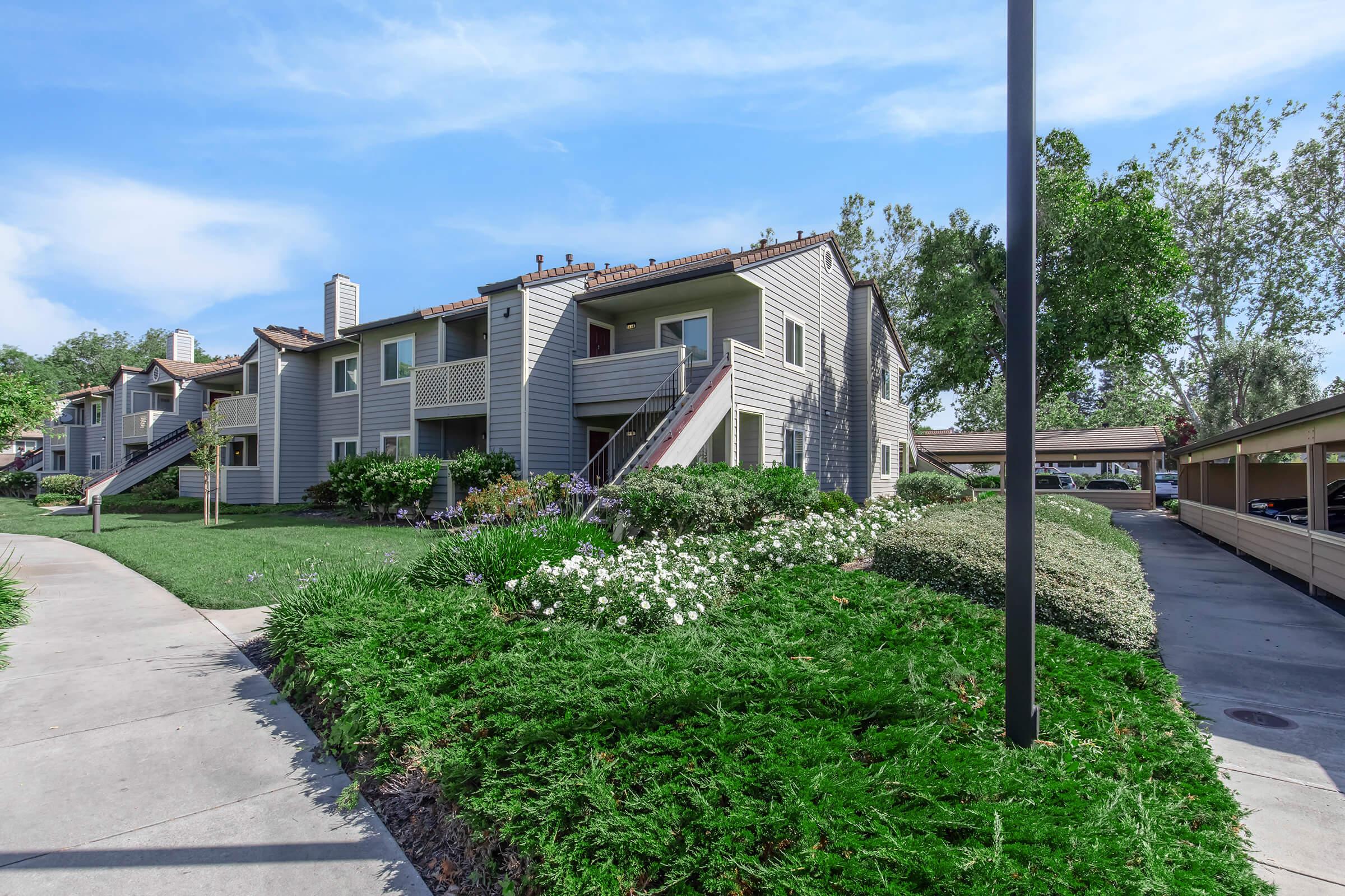 A landscaped apartment complex featuring two-story buildings with balconies, surrounded by green grass and flowering bushes. A walkway leads through the garden area, with a covered parking space visible on the right side. Trees provide shade in the background under a clear blue sky.