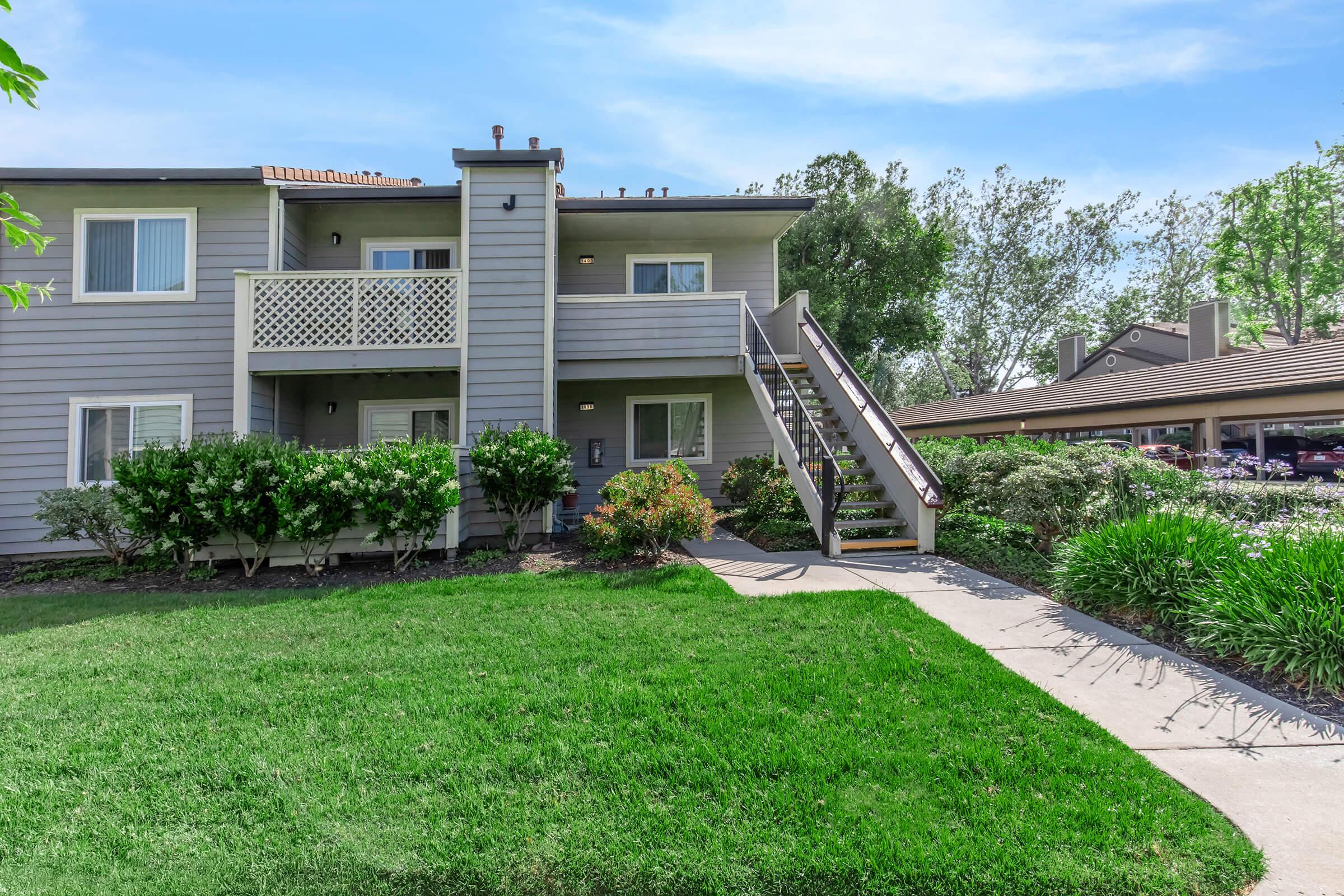 Two-story apartment building with a staircase leading to the upper level, surrounded by shrubs and greenery. A concrete pathway leads to the entrance, and a covered parking area is visible in the background, along with trees and blue sky.