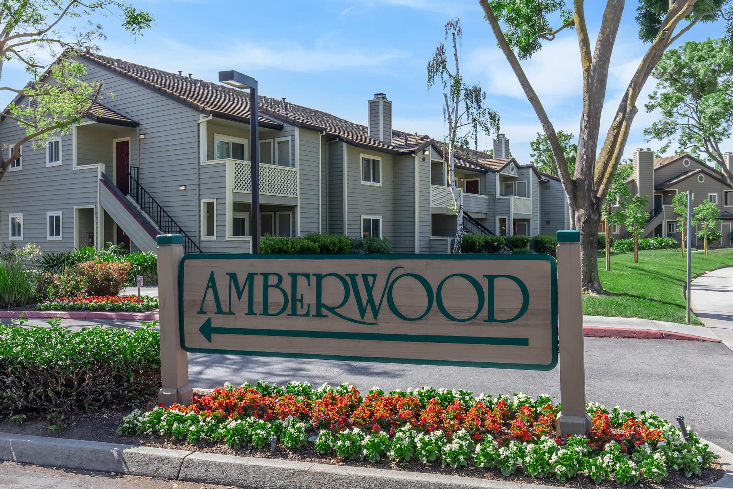 Sign for Amberwood community with landscaped flowers in the foreground, surrounded by apartment buildings and green trees in a residential setting.