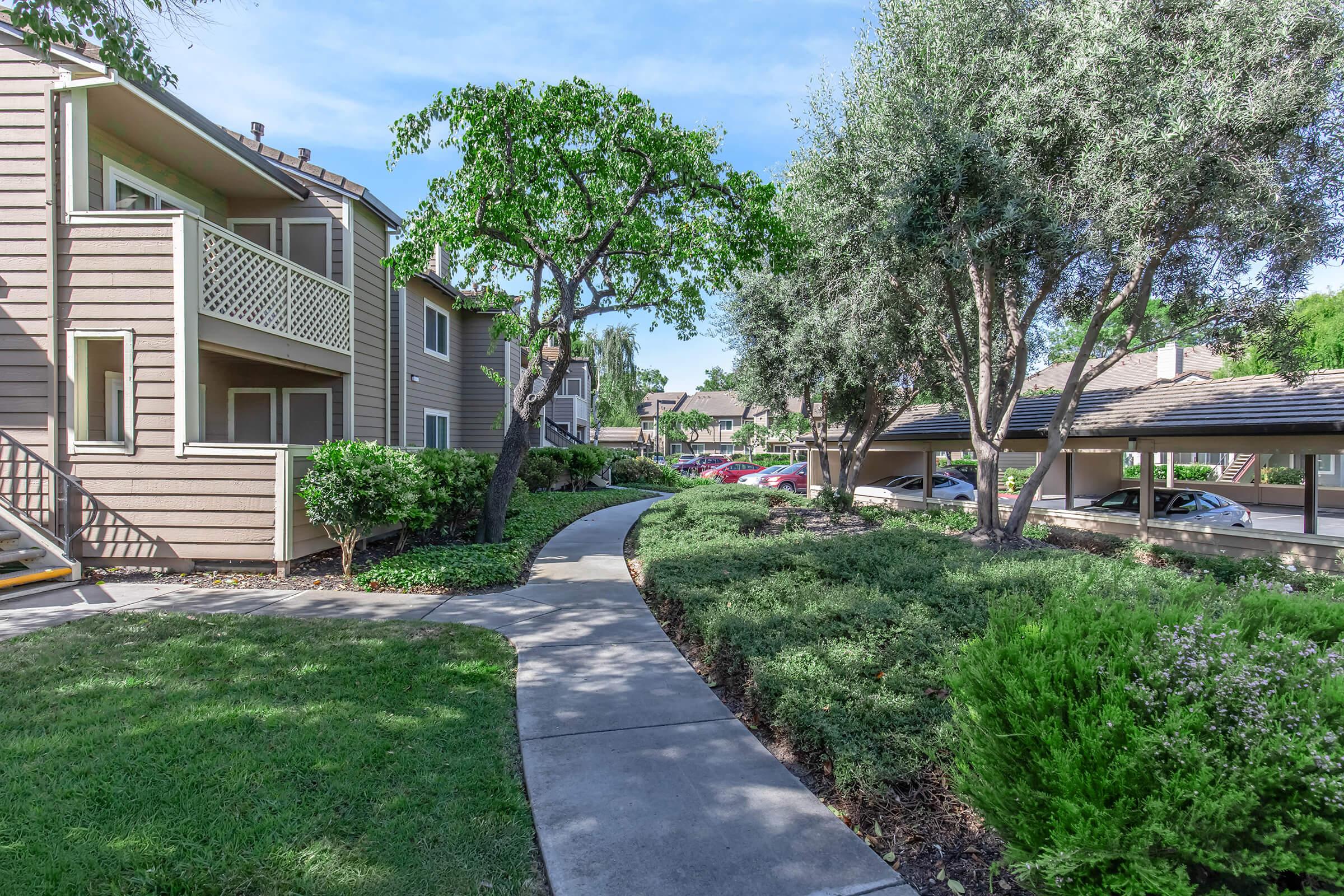A well-maintained landscaped path winding through an apartment complex, lined with greenery and trees. On one side, there are two-story buildings with balconies, and on the other, covered parking spaces are visible. The scene is bright and sunny, suggesting a pleasant environment.