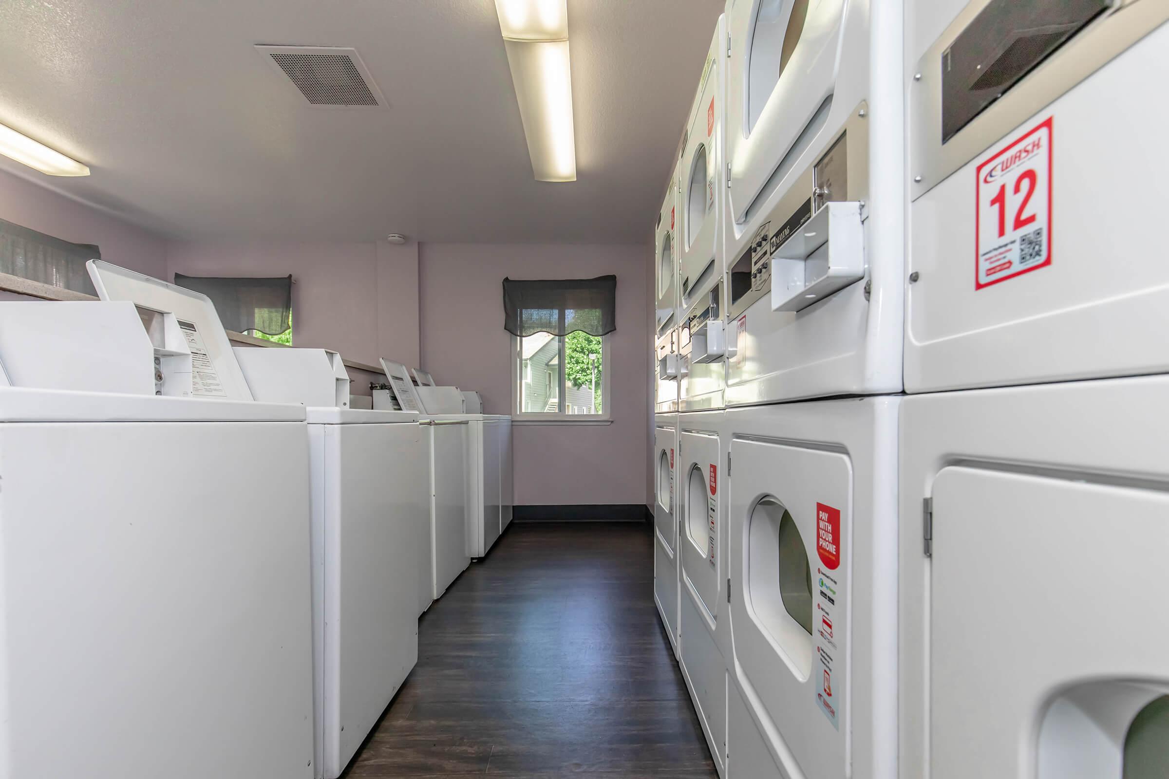 A laundry room featuring multiple white washing machines and dryers lined up against the walls. The space has a light-colored interior with overhead lighting, a window letting in natural light, and a clean, modern aesthetic.