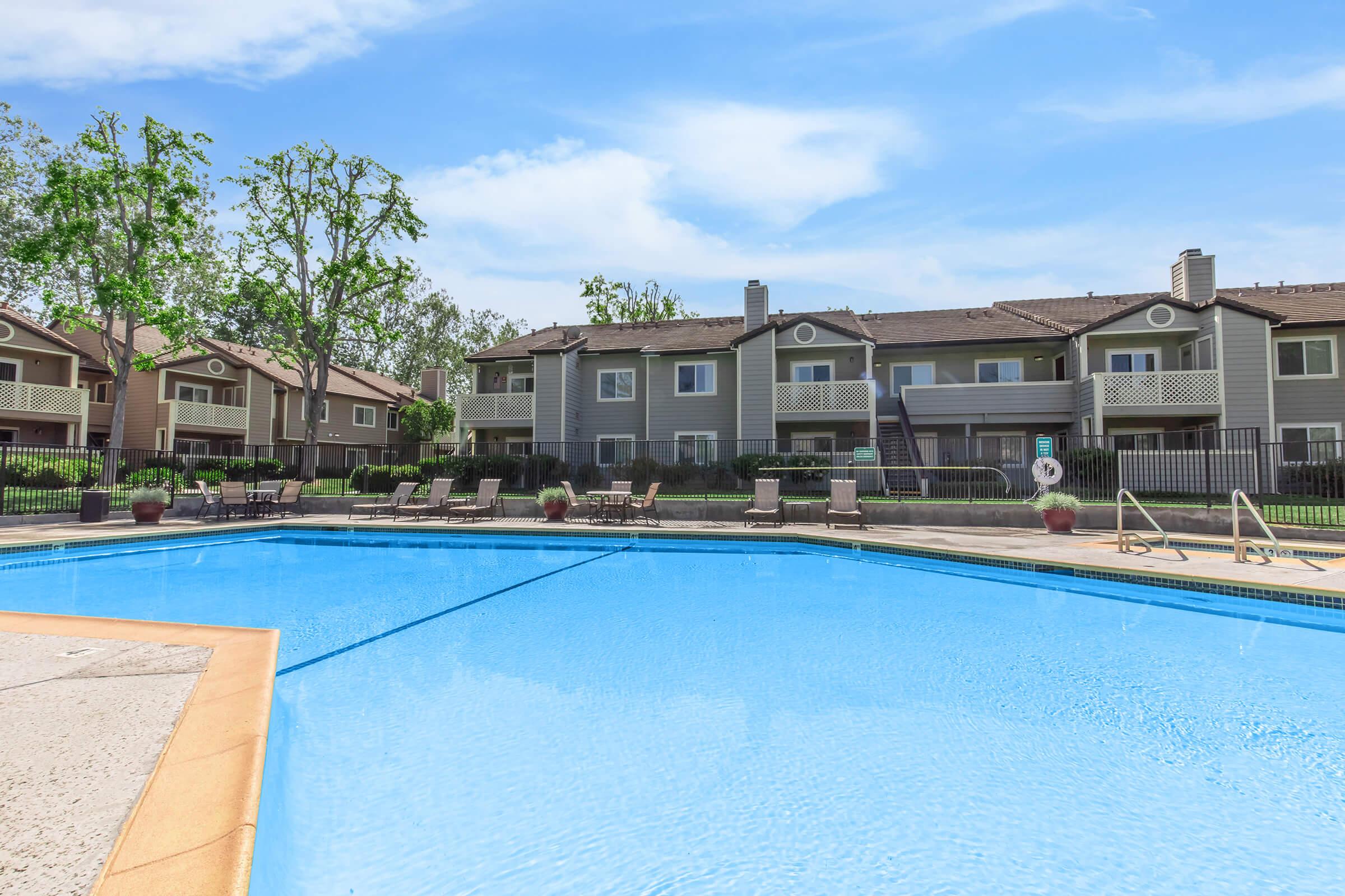 A clear blue swimming pool surrounded by lounge chairs and potted plants, with two-story residential buildings in the background. The scene is set in a green, landscaped area under a bright blue sky with scattered clouds.