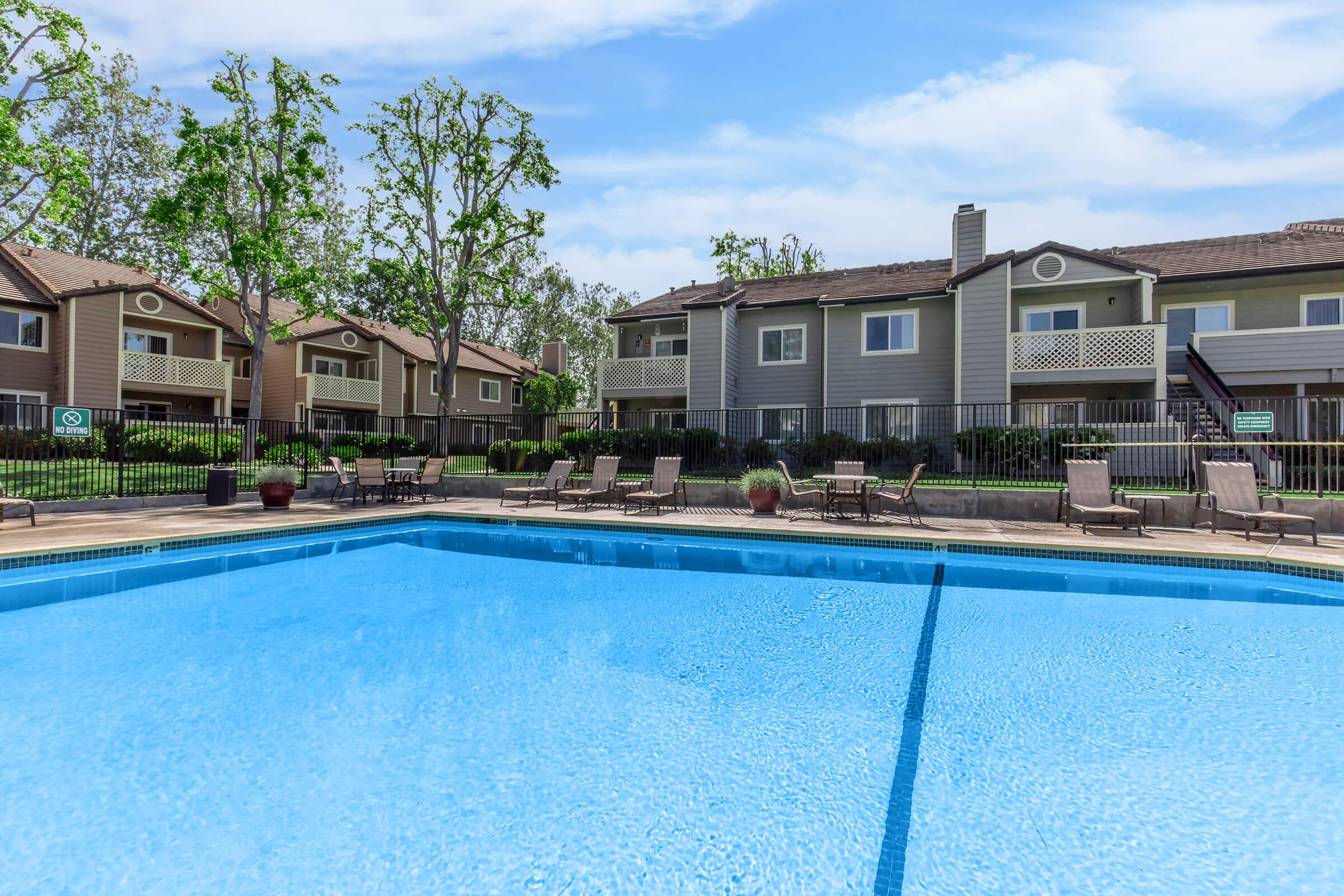 A clear blue swimming pool in the foreground with lounge chairs arranged around it. In the background, there are two-story apartment buildings with balconies, surrounded by lush green trees and landscaped gardens. The sky is bright and partly cloudy, creating a refreshing atmosphere.