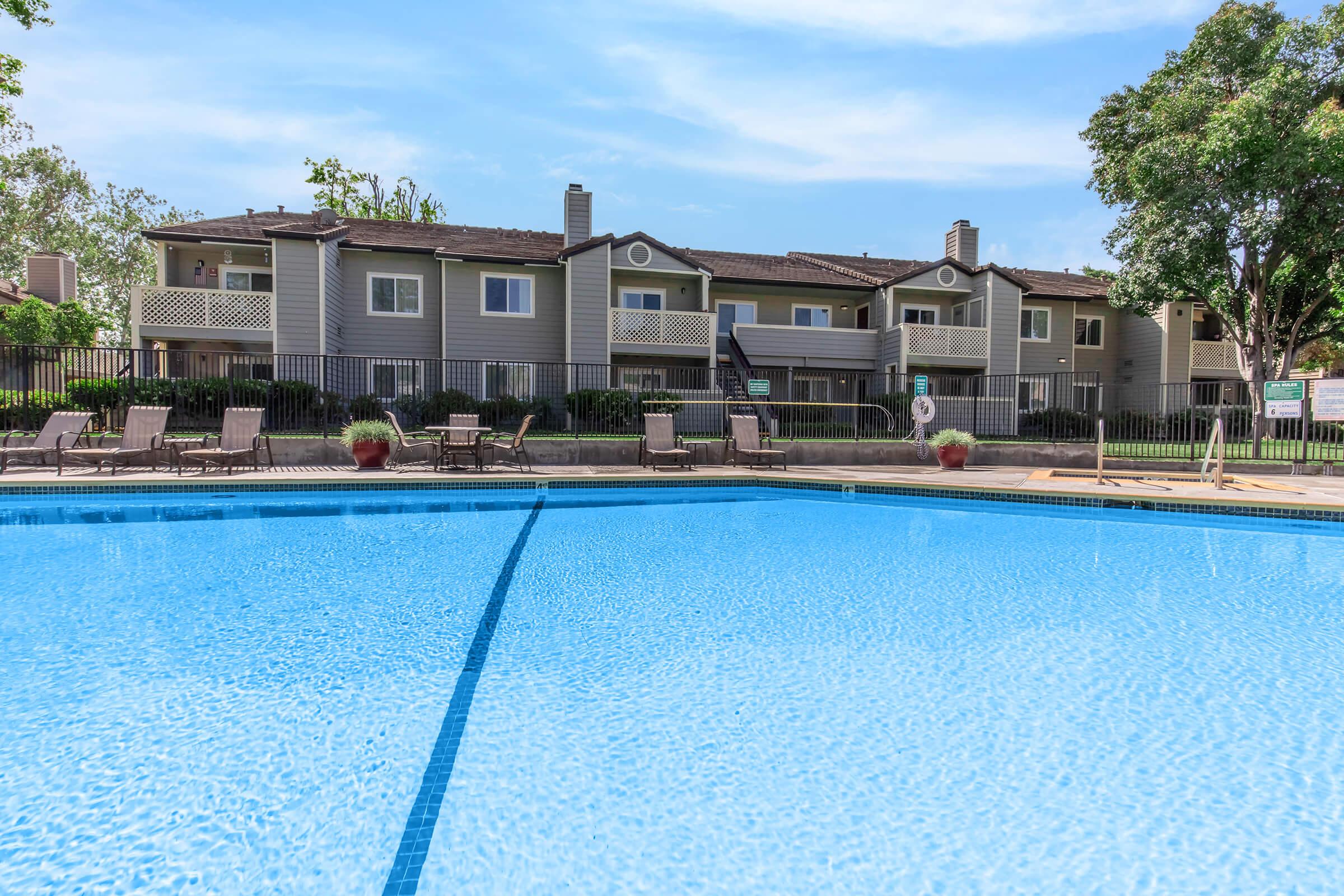A view of a swimming pool with crystal-clear blue water in the foreground, surrounded by lounge chairs. In the background, there is an apartment complex with balconies and green trees. The scene is set on a bright, sunny day, highlighting a serene and inviting atmosphere.