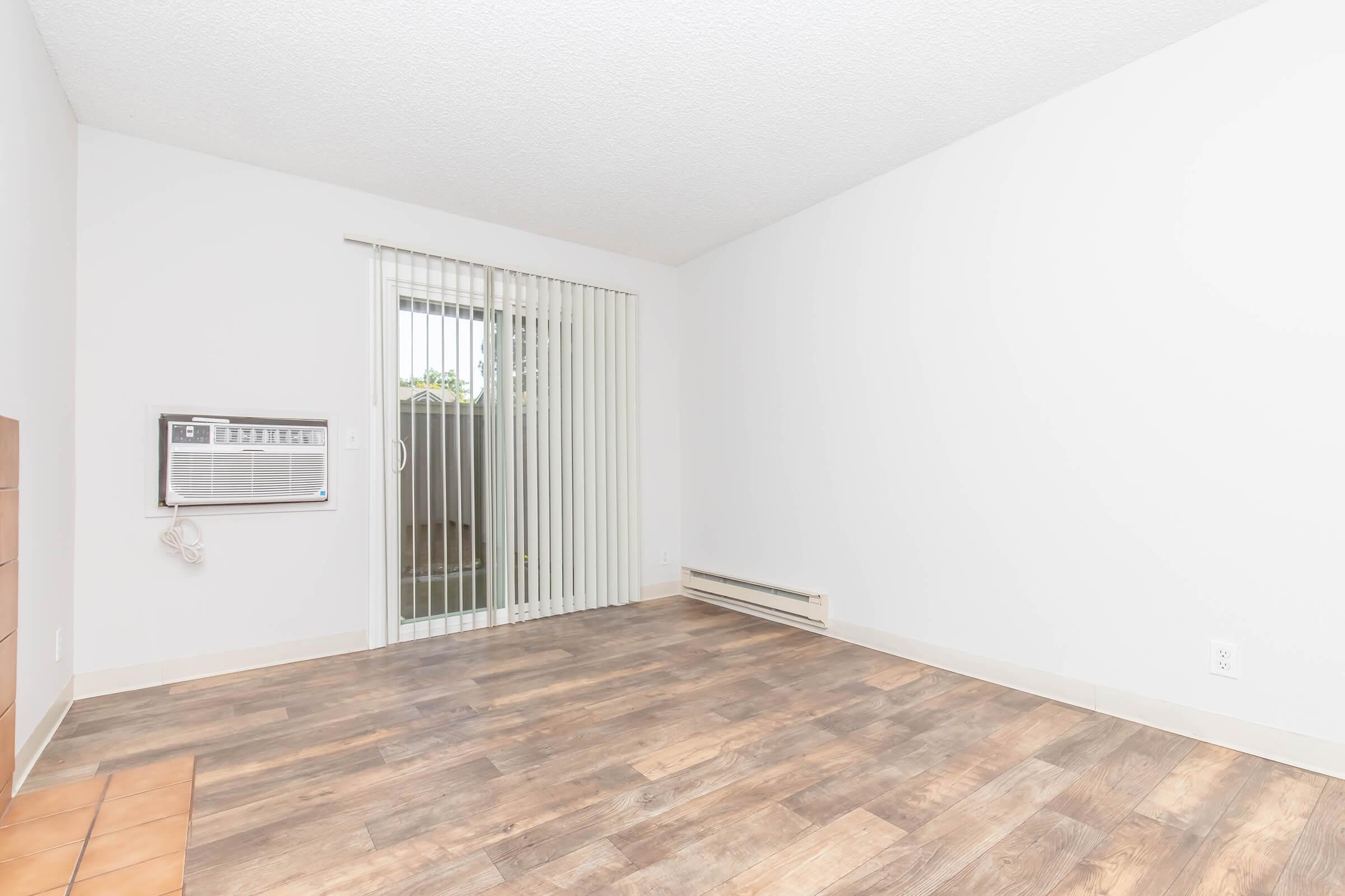 A spacious, empty room with light-colored walls and wooden laminate flooring. An air conditioning unit is installed on the left wall, and there is a sliding glass door with vertical blinds leading outside. The room is well-lit, creating an open and airy atmosphere.