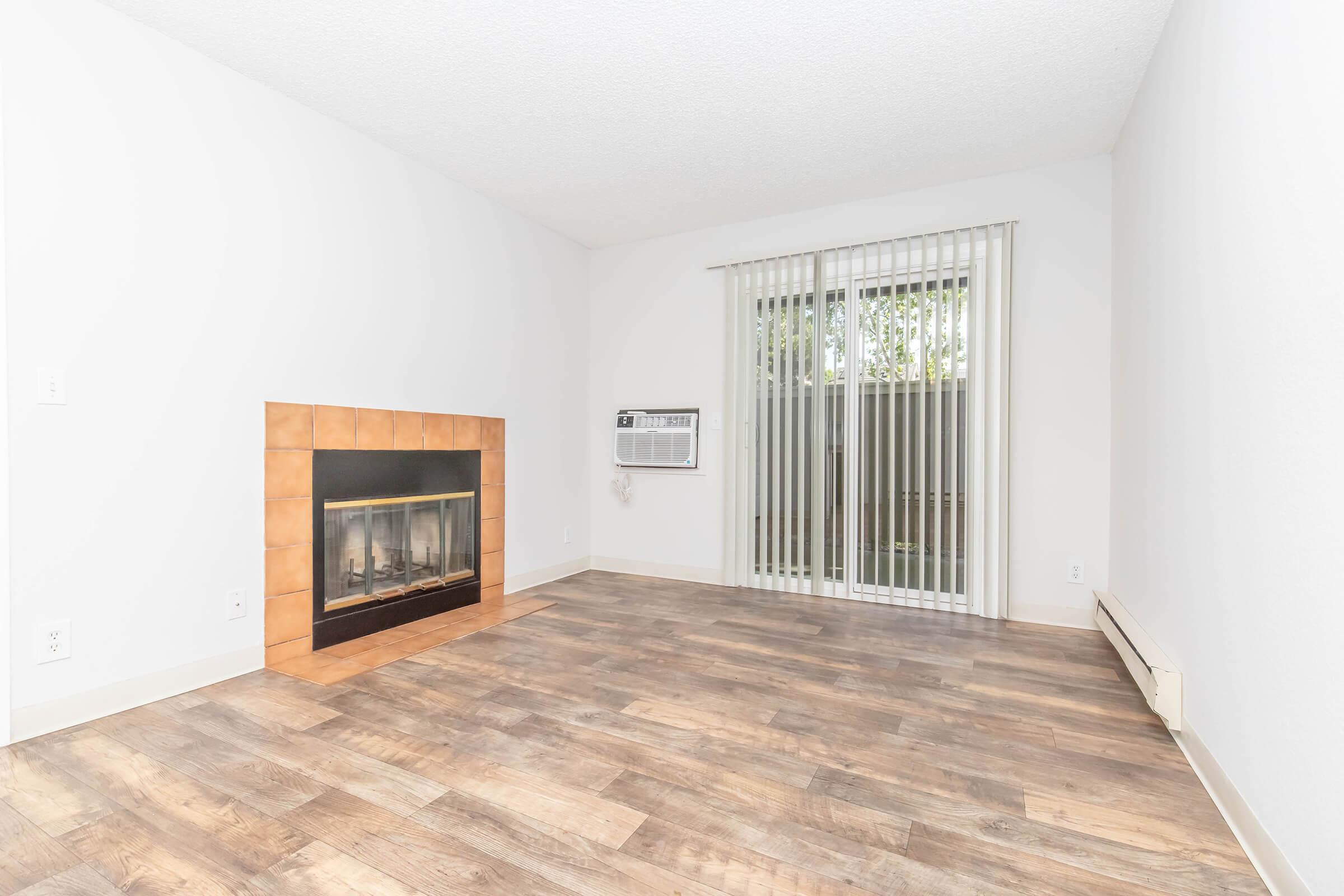 Empty room featuring a fireplace with a tiled surround, large windows with vertical blinds, and a wall-mounted air conditioning unit. The flooring is a warm-toned wood laminate, and the walls are painted white, creating a bright and airy atmosphere.