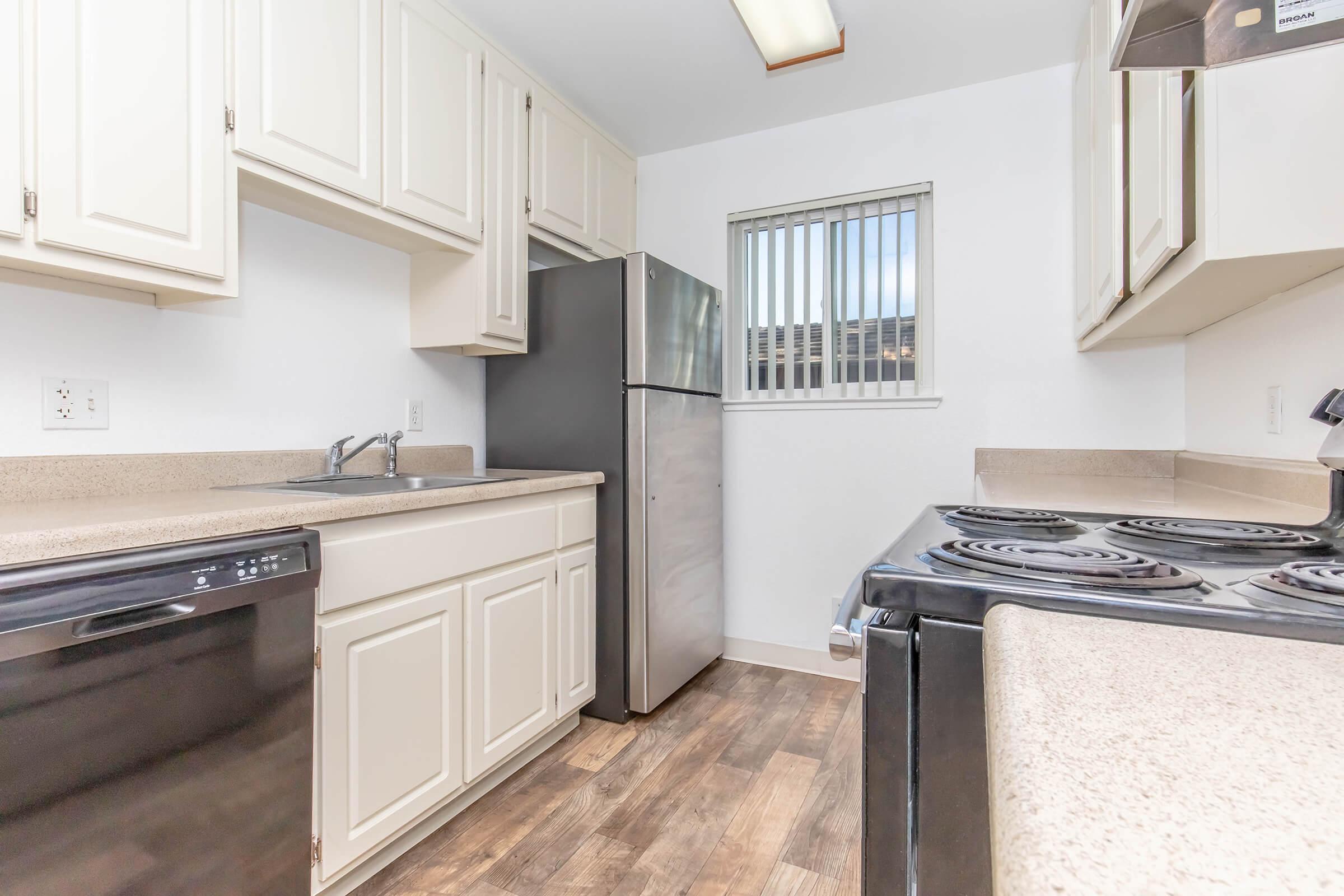 A modern kitchen featuring white cabinets, a stainless steel refrigerator, an electric stovetop, and a black dishwasher. The countertop is light-colored, and a window with vertical blinds allows natural light. The flooring is a wood-like material, creating a warm atmosphere.