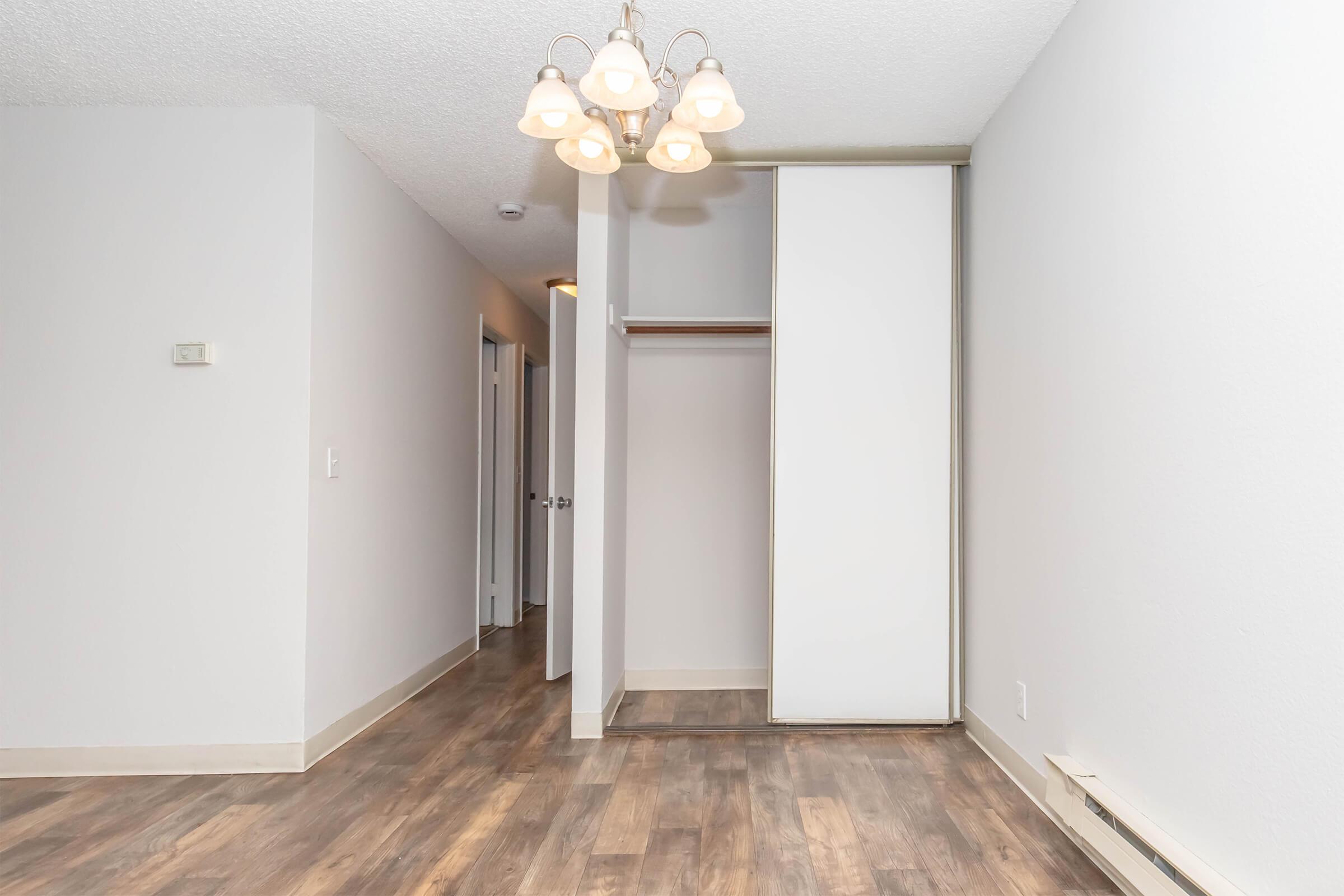 A well-lit interior space featuring light gray walls and wooden flooring. A chandelier with five light bulbs hangs from the ceiling. On the left, a closet with sliding doors is visible, and a doorway leads to another room in the background. The overall area appears clean and spacious.