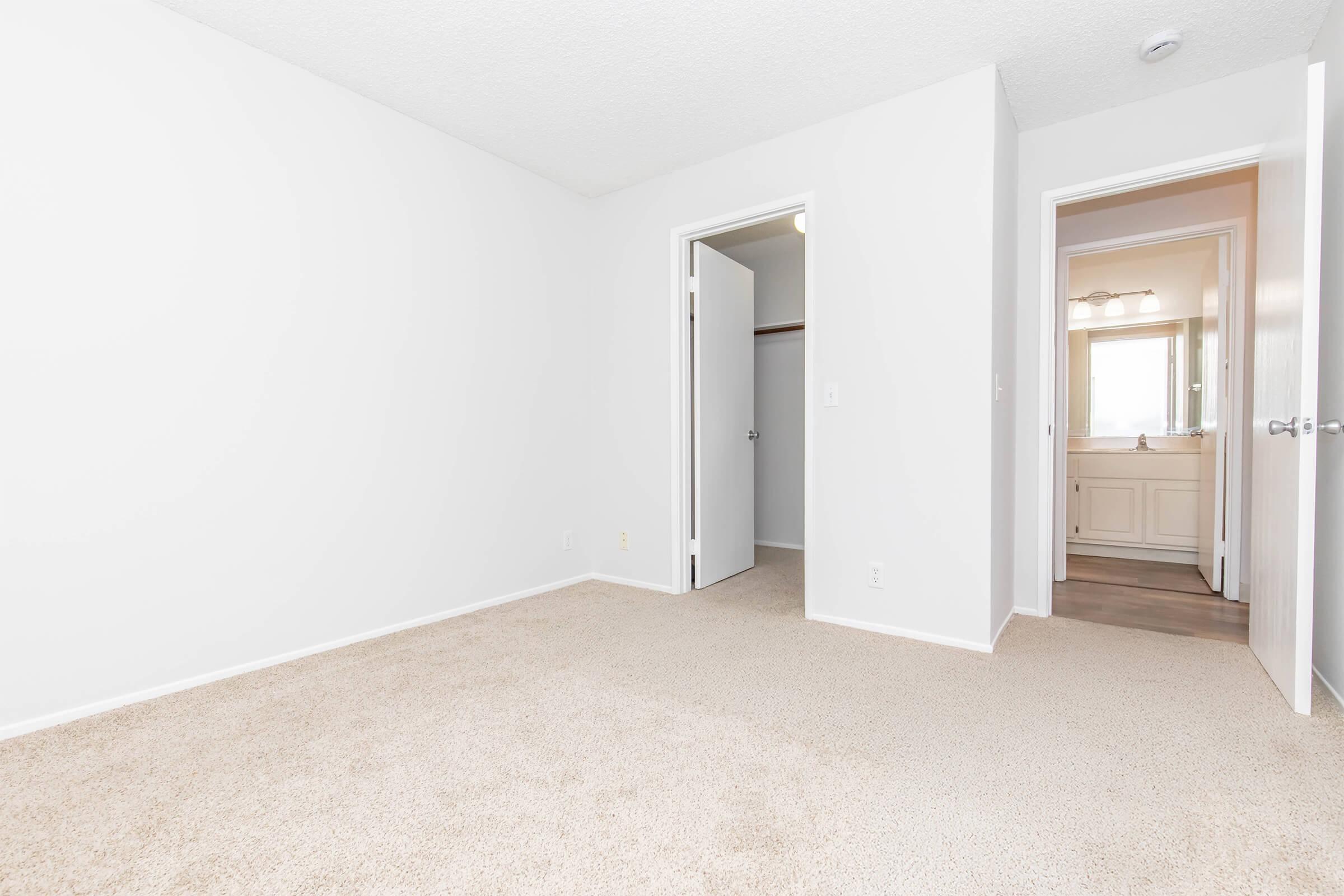 Empty room featuring beige carpet and plain white walls. Two doorways are visible: one leading to a closet and another to a bathroom or hallway. Natural light is coming from the entryway, creating a bright and spacious atmosphere.