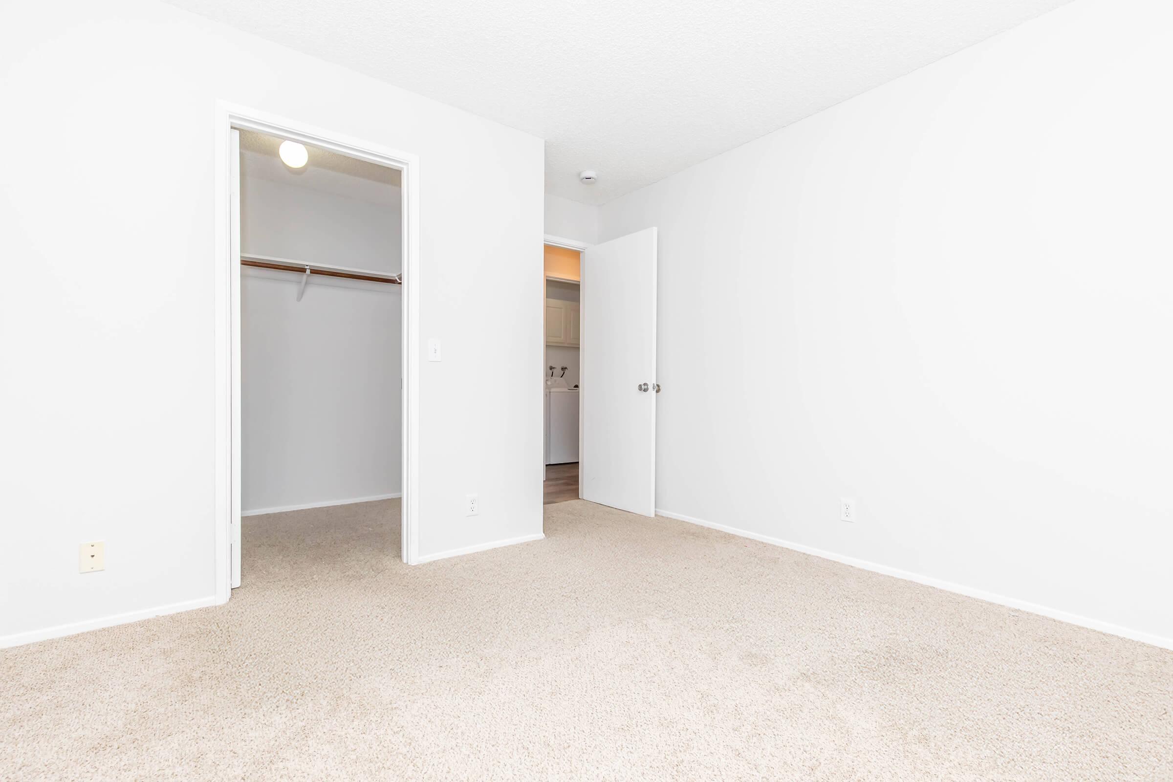 Empty room with light-colored walls and carpet, featuring an open door on the left leading to a closet and a closed white door on the right. The space is bright and minimalistic, showcasing a simple design suitable for various uses.