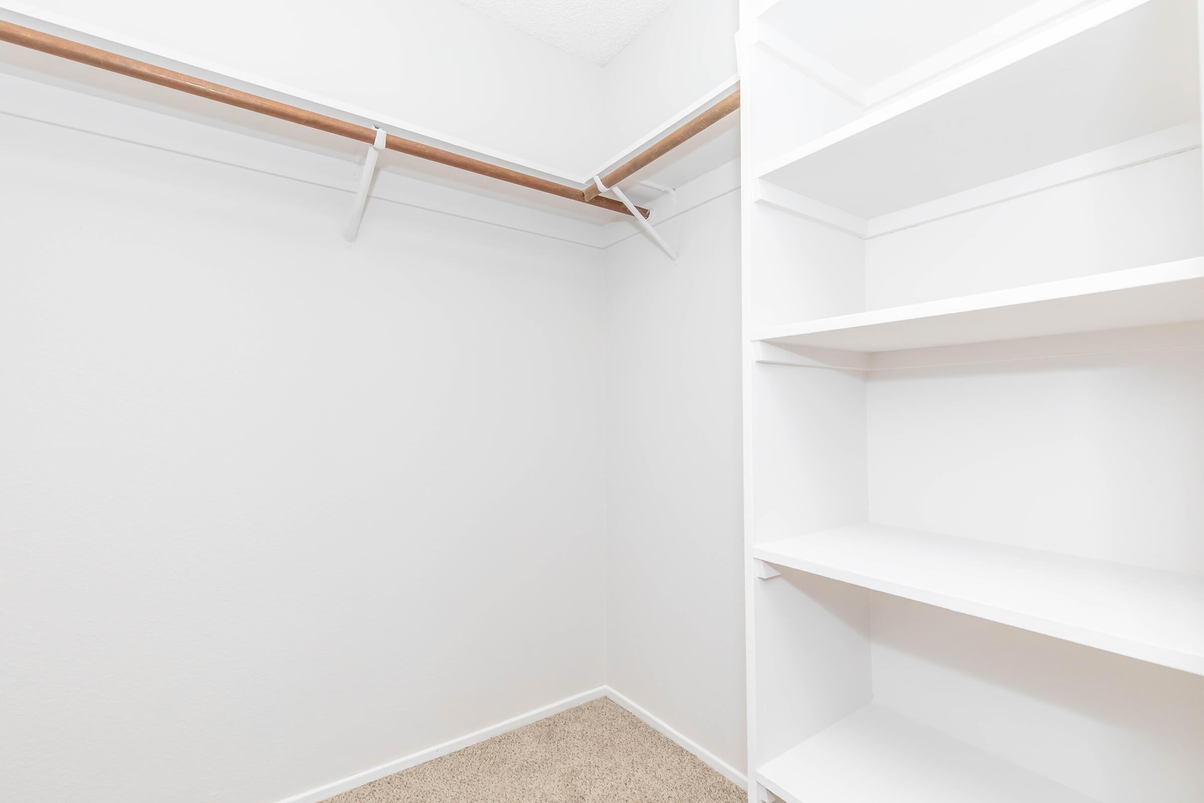 A clean, empty closet with a white interior. It features a single clothes rod along the top and a set of built-in shelves on one side, all against a light background. The floor is covered with beige carpeting.